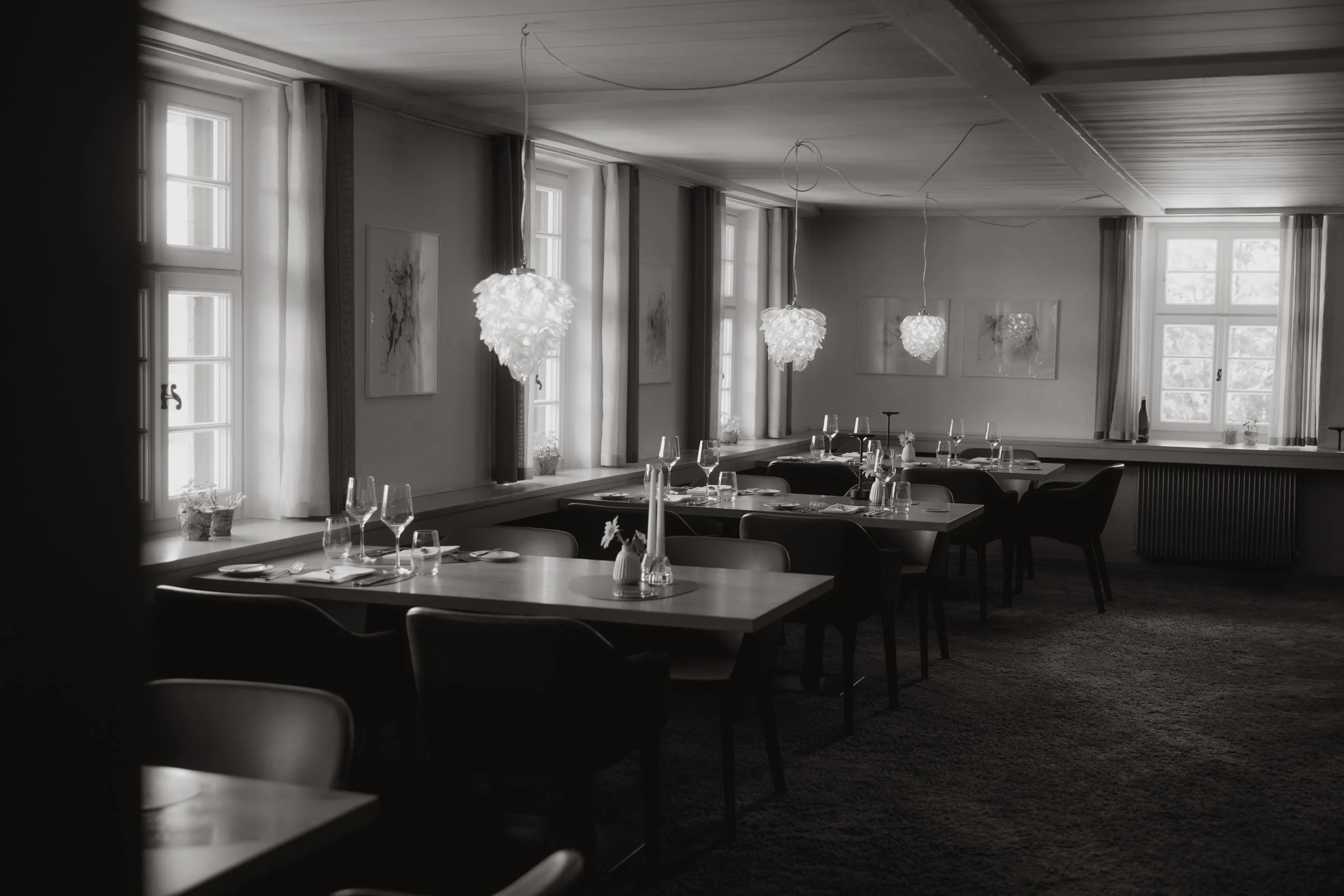 Zeitloser Speiseraum im Restaurant Hohenkarpfen mit mehreren Tischen, Gläsern, Tellern und Kerzen, beleuchtet durch natürliche Lichtquellen und elegante Lampen, mit modernen Kunstwerken an den Wänden. Fotografiert von Pia Heim an einem Event.