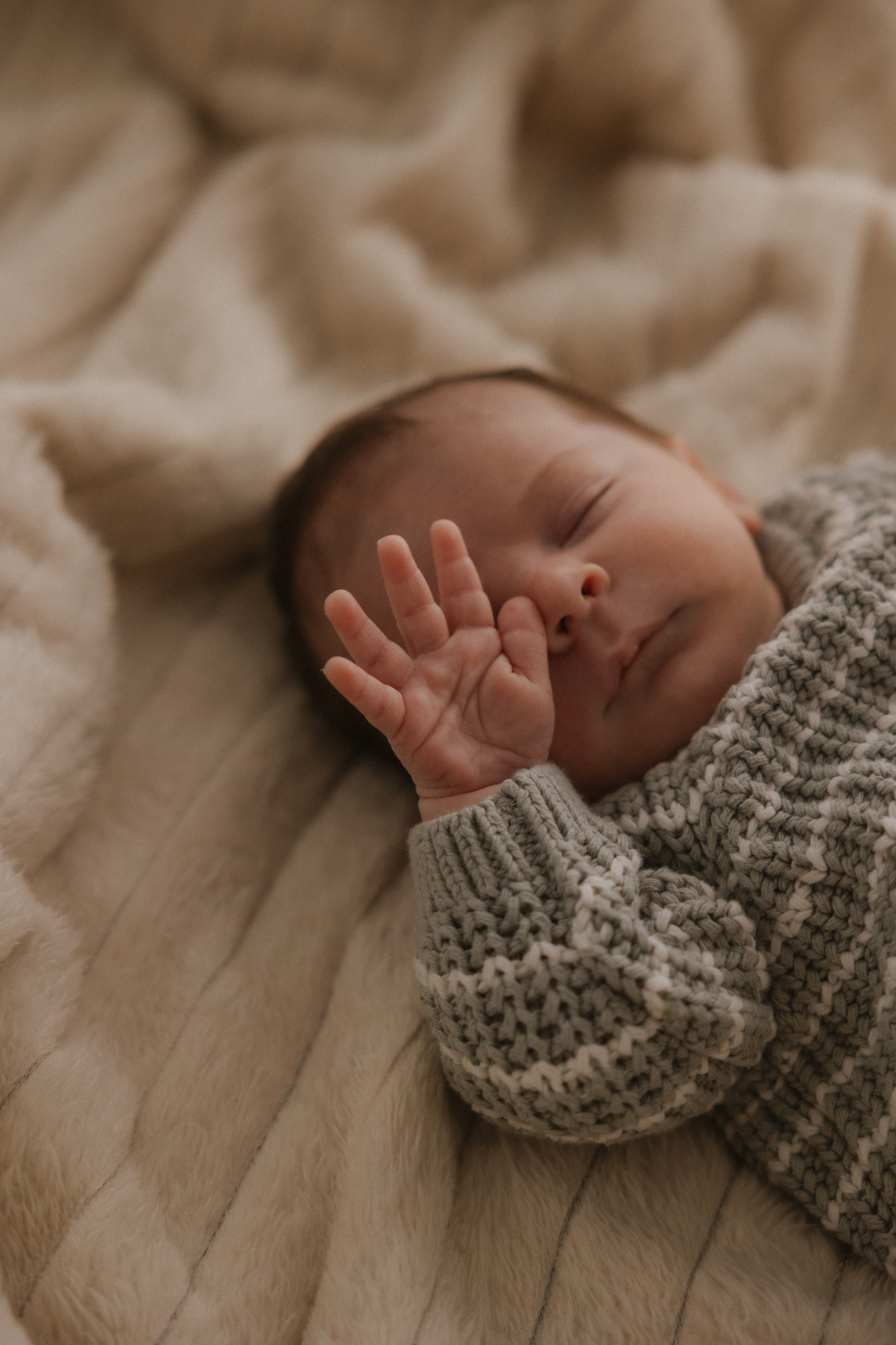 Ein schlafender Säugling mit geschlossenen Augen, der auf einem weichen, beigen Fell liegt, trägt einen grauen, gestrickten Pullover. Das Baby zeigt mit der Hand eine Geste, vermutlich ein kleines Handzeichen.