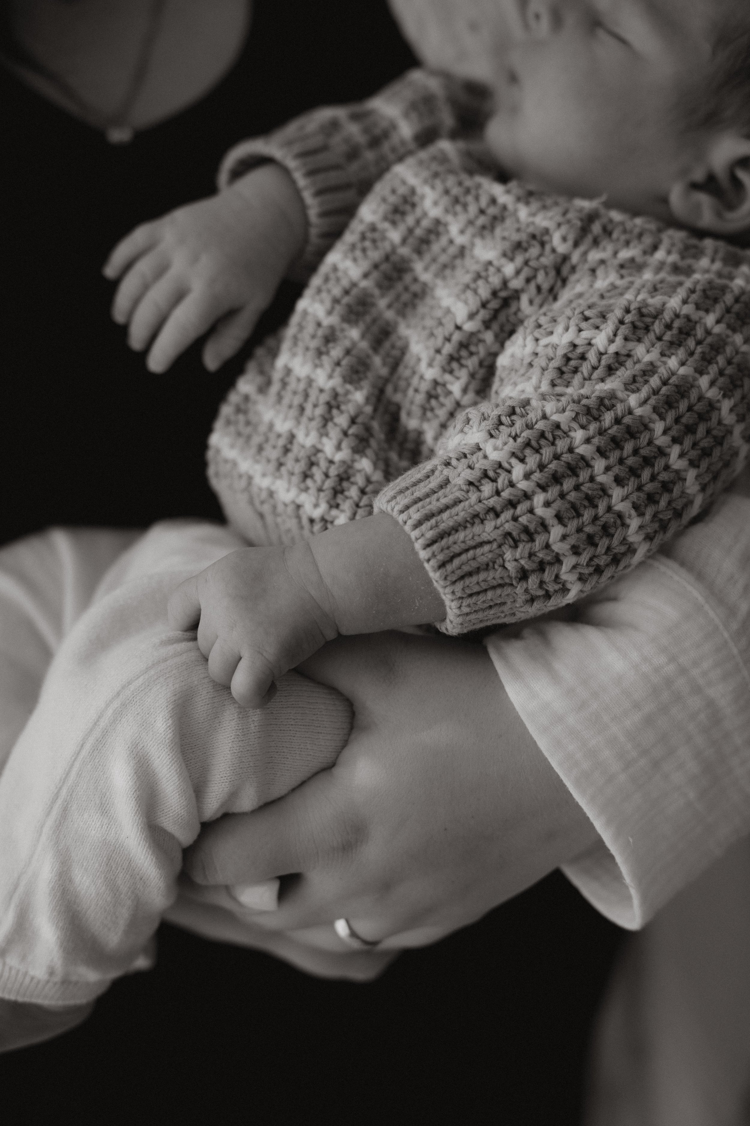 Nahaufnahme eines Erwachsenen, der ein Kind hält. Das Kind trägt eine gestreifte Strickjacke und eine langärmelige Kleidung, die Elternfigur trägt einen hellen Hemdkragen. Es ist in Schwarz-Weiß fotografiert.
