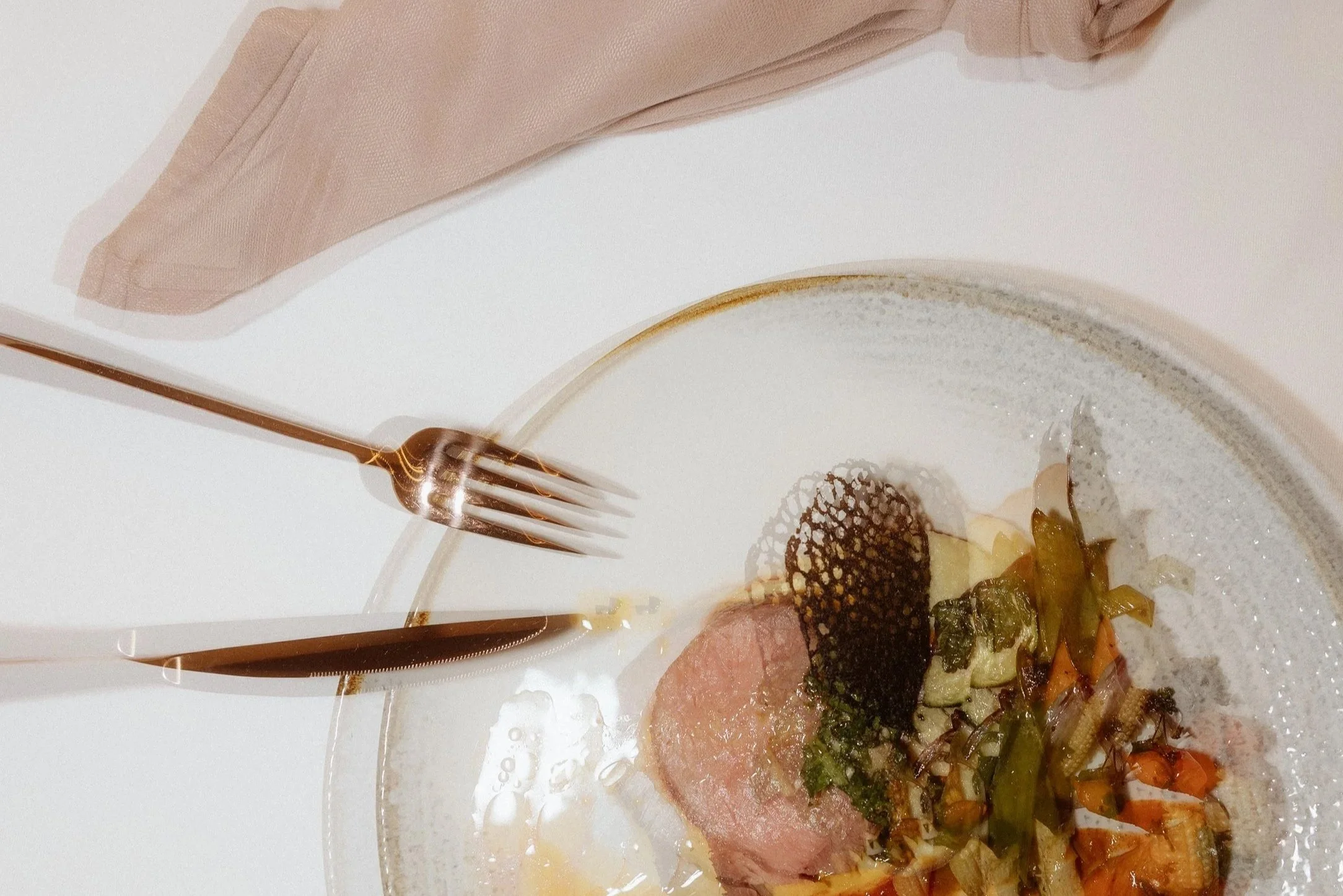Moderne Foodfotografie der Eventschneiderei mit gekochtem Bio-Gemüse, einer Scheibe Fleisch und Garnitur, Messer und Gabel auf weißem Tisch, mit beige Stoffserviette im Hintergrund.