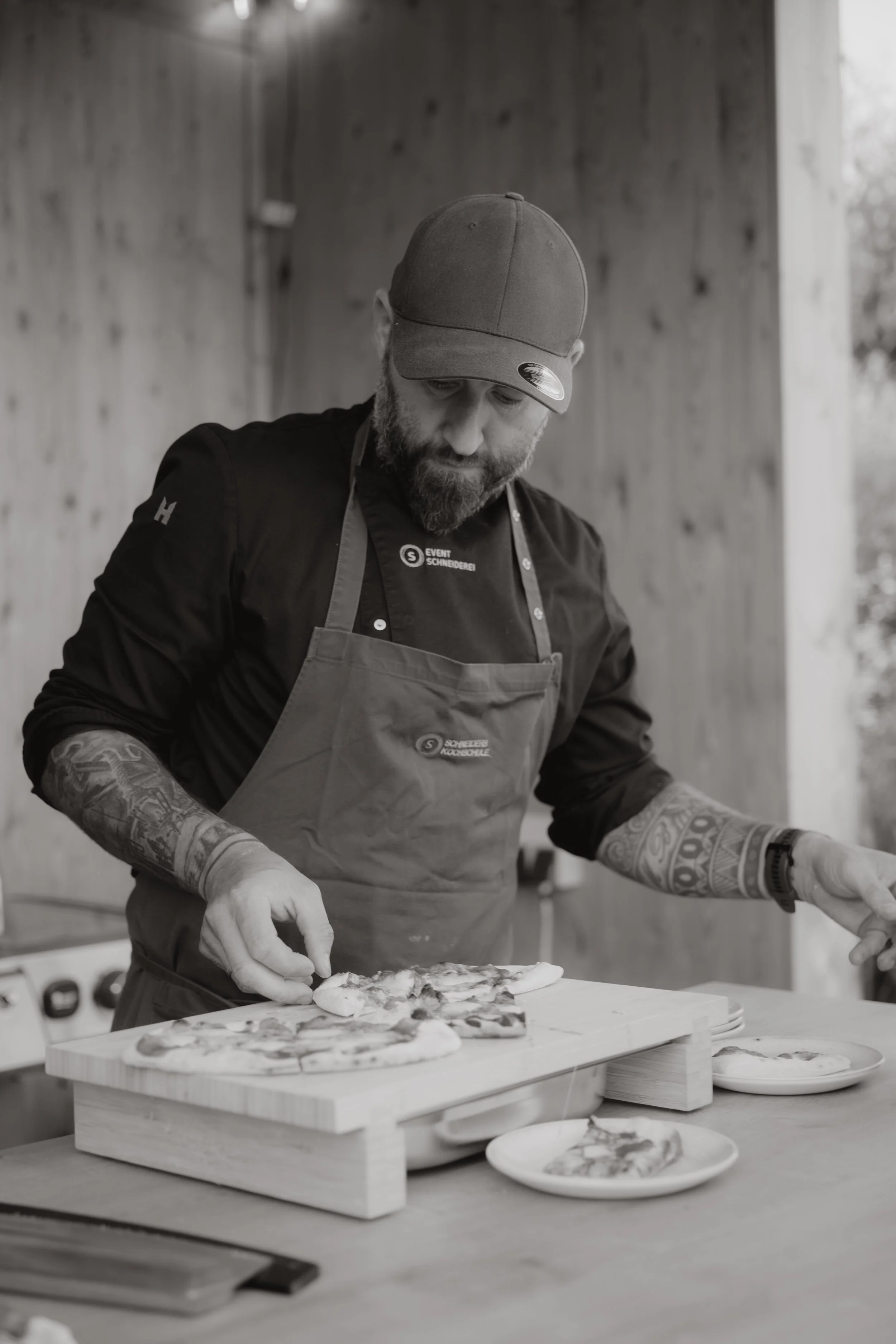 Ein Koch mit Tattoos auf den Armen, einer Schürze und Mütze bereitet Pizza in der Holzhütte der Eventschneiderei vor. Fotografische Begleitung der Open Kitchen Party durch Pia Lydia Heim. 