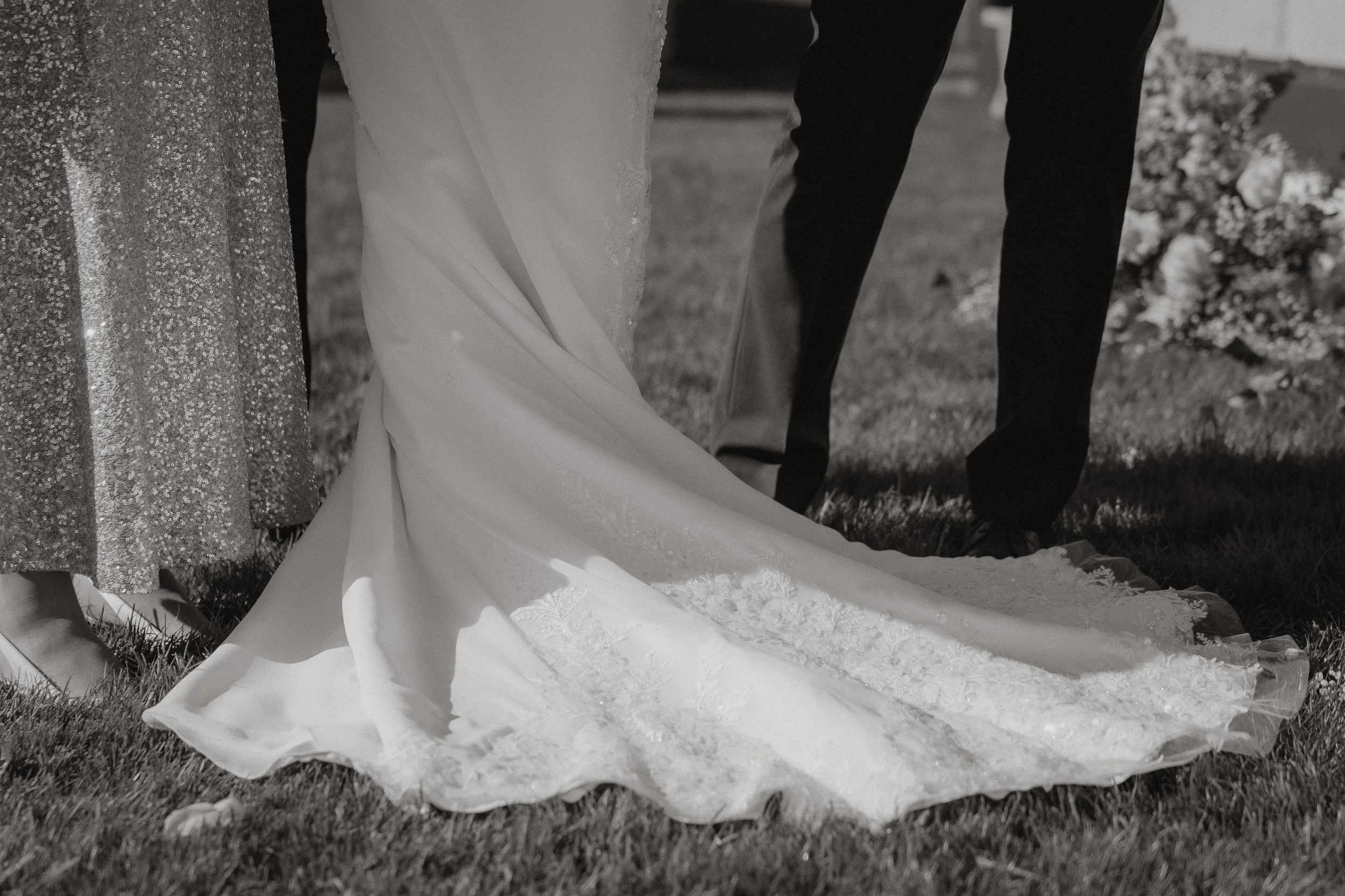 Detailaufnahme eines Brautkleids, eines Anzuges und eines Brautpaares bei einer Hochzeit im Freien, aufgenommen in Schwarz-Weiß.