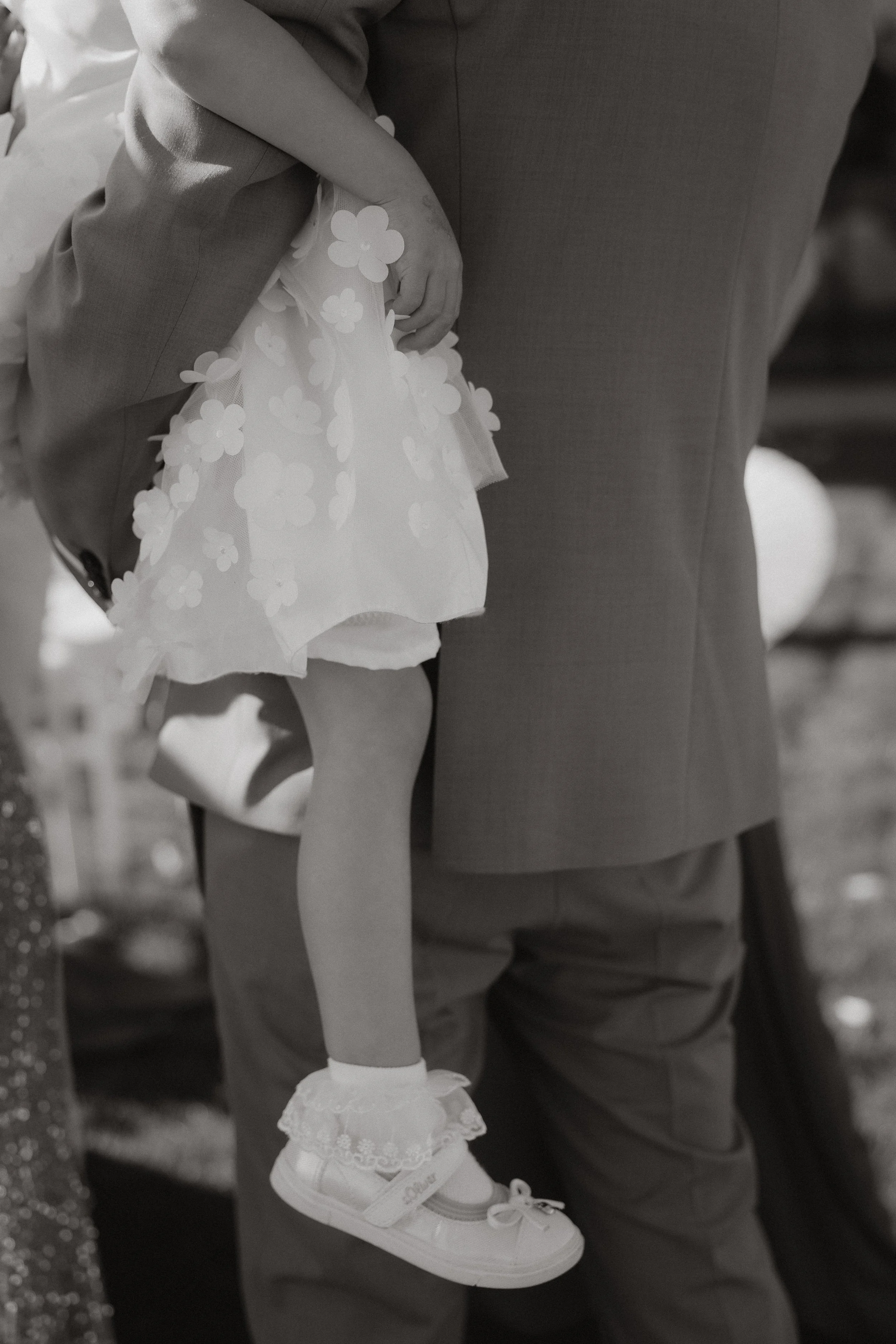 Ein kleines Mädchen wird auf einer Hochzeit  von ihrem Papa gehalten. Nur die Beine und ein Teil des Oberkörpers des Erwachsenen sind sichtbar. Das Mädchen trägt eine Blümchen-Party-Kleidung mit weißen Schuhen und Socken.