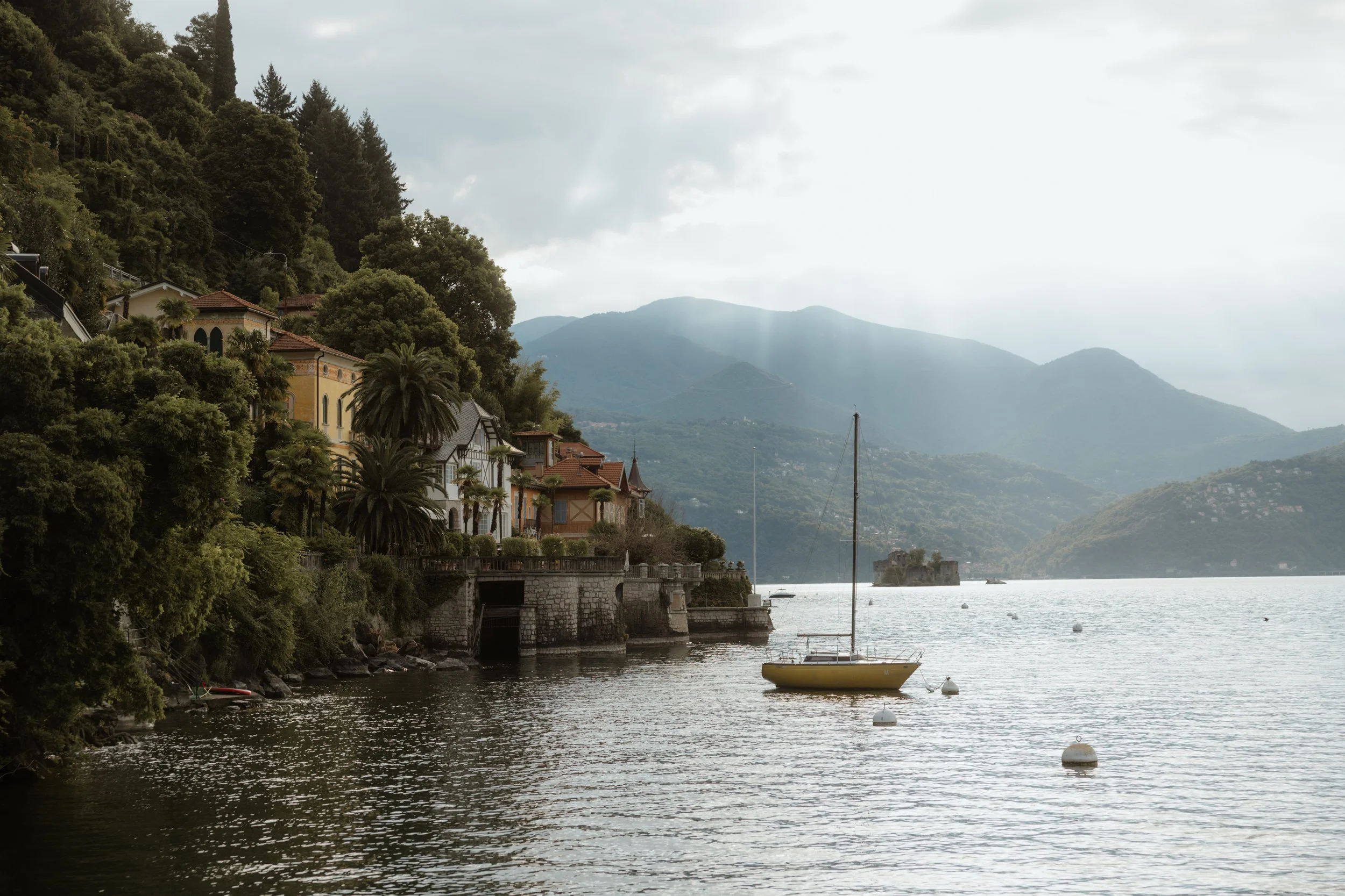 Blick auf einen See mit Segelbooten, umgeben von Häusern und grünen Hügeln, im Hintergrund Berge und bewölkter Himmel. Destination Wedding mit Weddingphotographer Pia Lydia Heim.