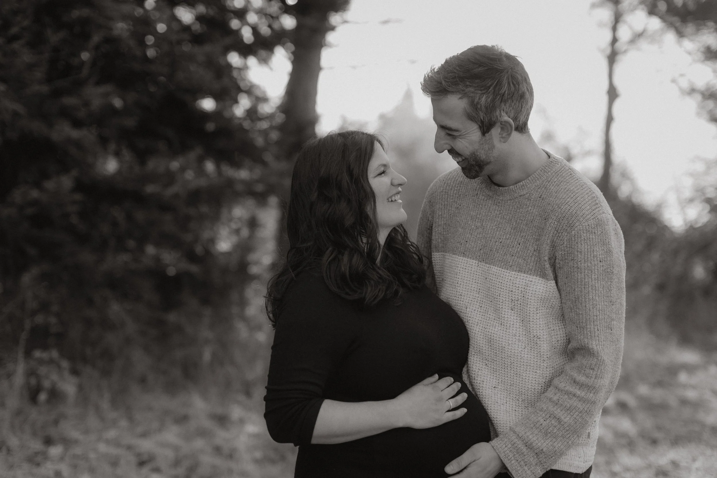 Ein lächelndes Paar, eine schwangere Frau und ein Mann, stehen draußen in der Natur und schauen sich liebevoll an.