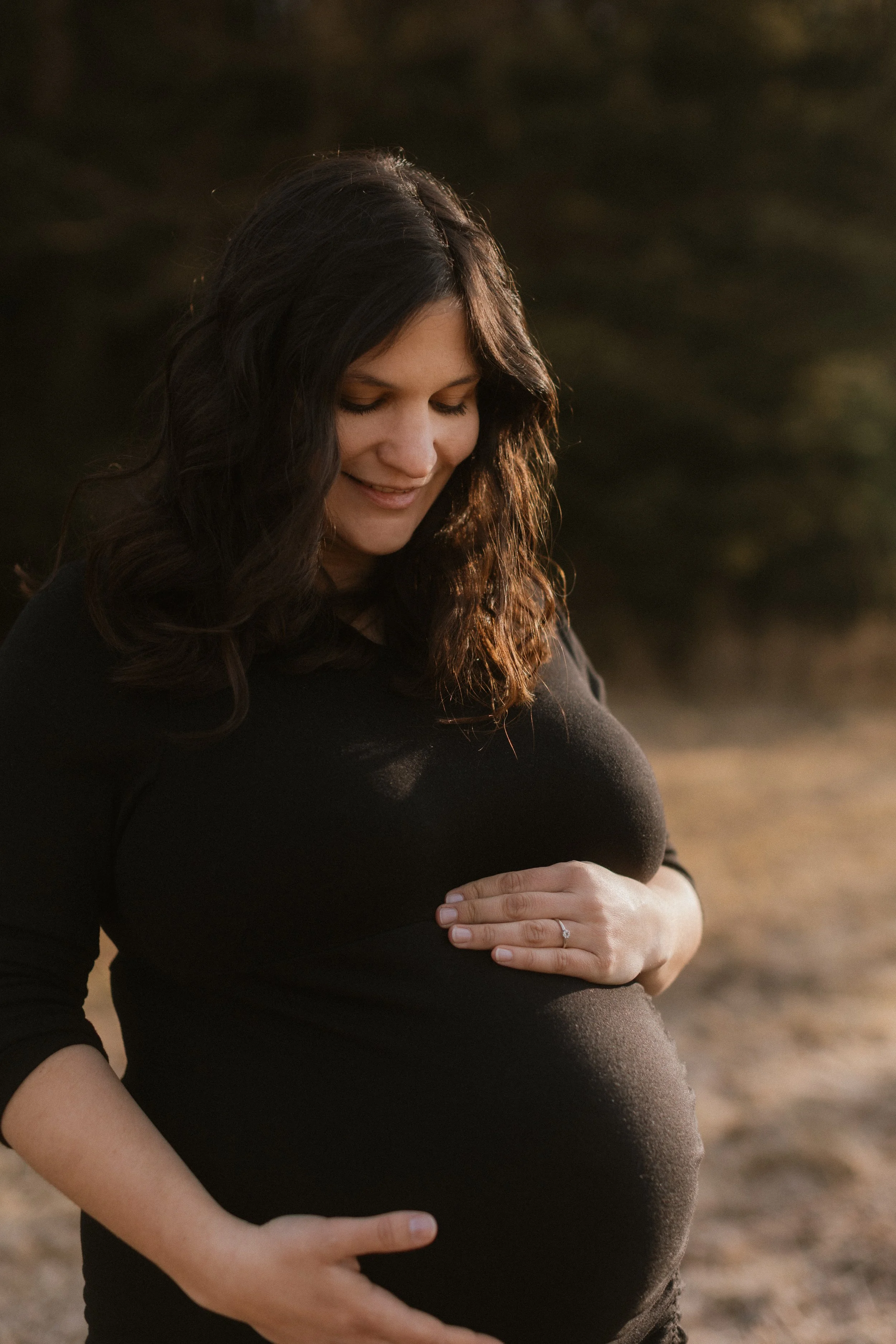 Schwangere Frau in einem schwarzen Kleid hält ihren Babybauch mit beiden Händen, lächelt und blickt nach unten, im Freien bei Sonnenuntergang.