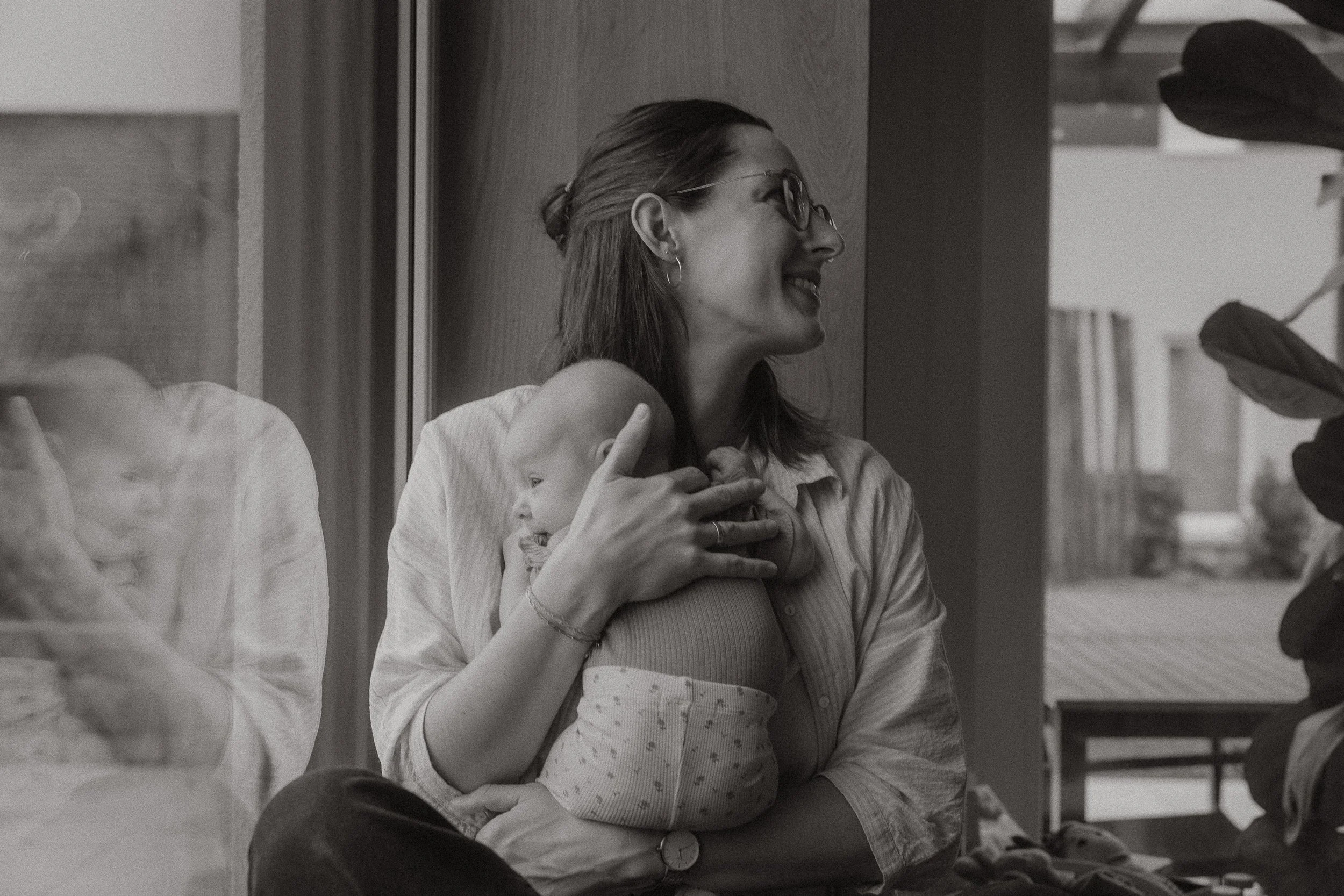 Eine Frau hält ein Baby auf dem Schoß, während sie lachend nach rechts schaut. Der Raum ist hell und modern, mit Holzpanel an der Wand und einer großen Fensterfront im Hintergrund.