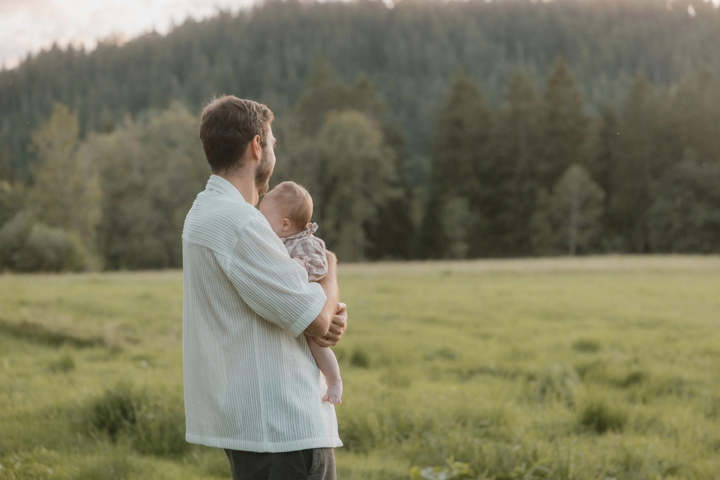 Ein Mann hält ein Baby in den Armen in einer grünen Wiese mit einem Wald im Hintergrund. Es ist vermutlich Sonnenuntergang, da das Licht warm ist.