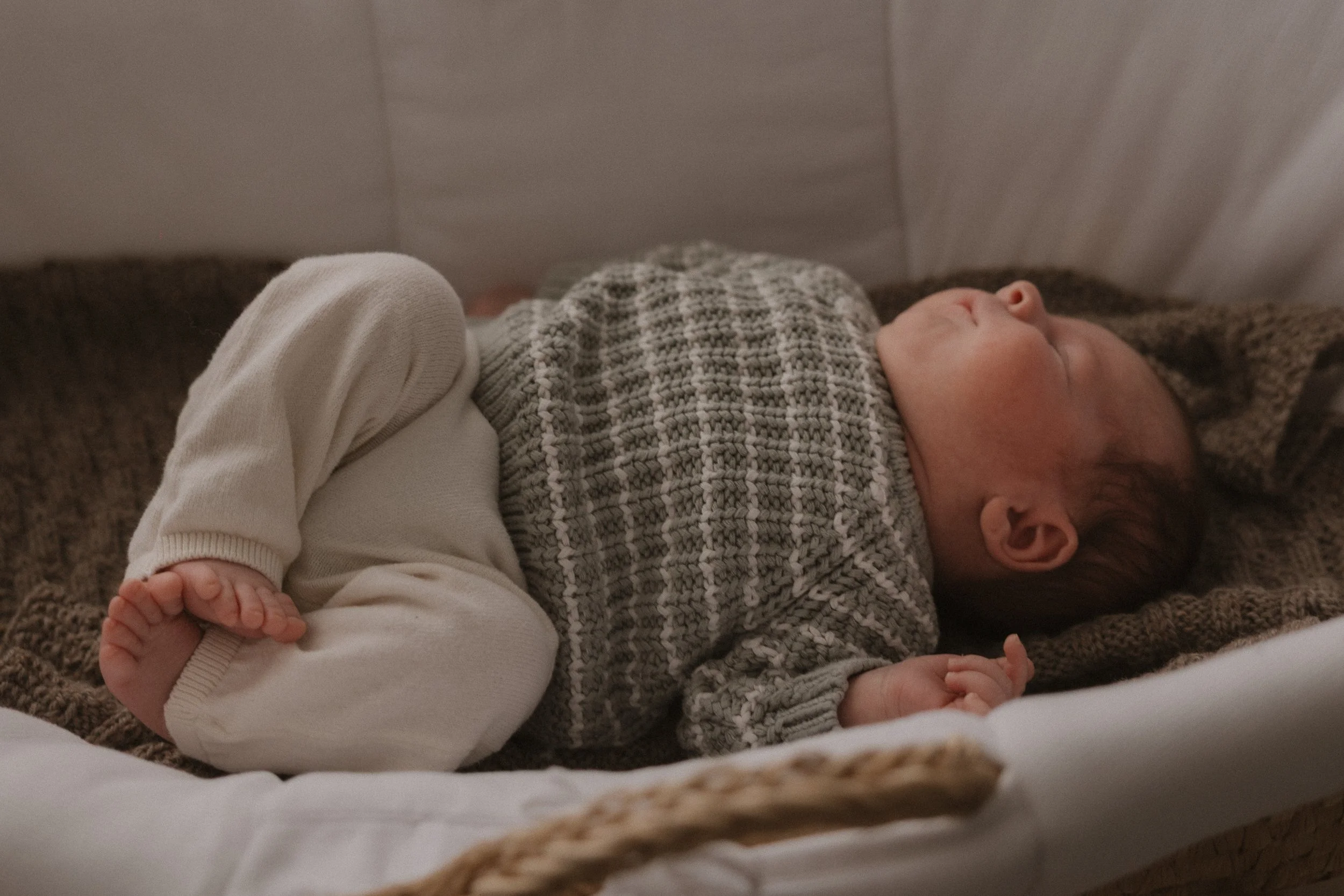 Ein schlafender Säugling liegt auf einer Decke in einem Bett, eingekleidet in einen grauen Pullover und hellen Babysöckchen.