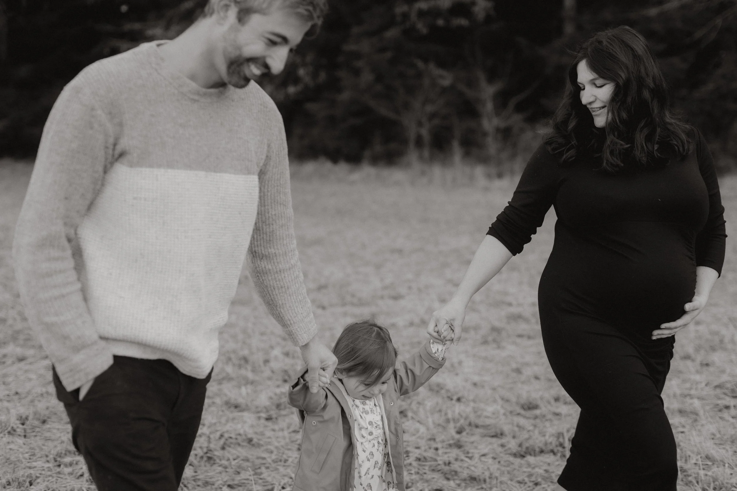 Ein Mann und eine Frau halten die Hände eines kleinen Mädchens, während sie im Freien spazieren. Die Frau ist schwanger.