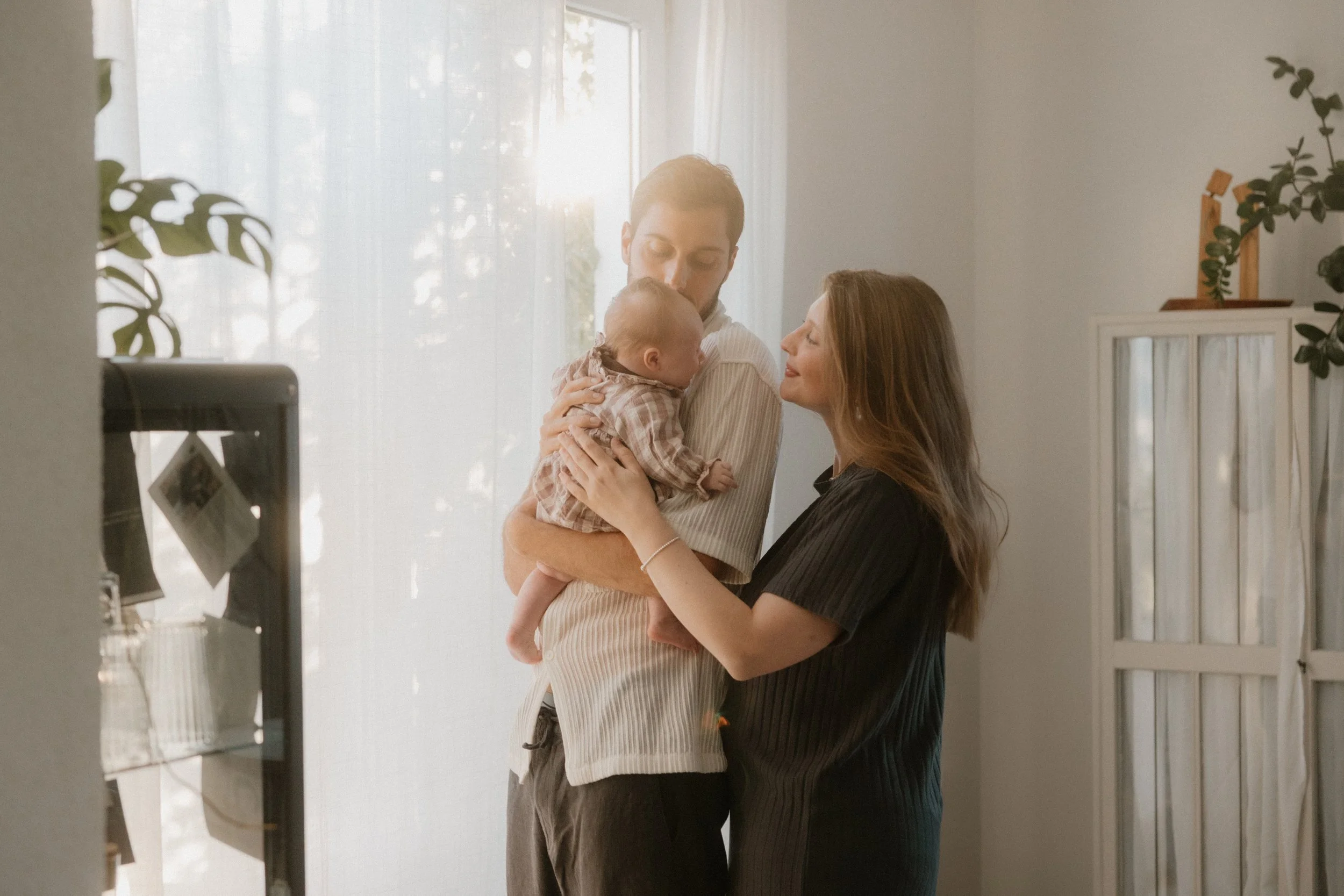 Ein Mann und eine Frau halten ein Baby in einem hellen Raum mit viel natürlichem Licht.