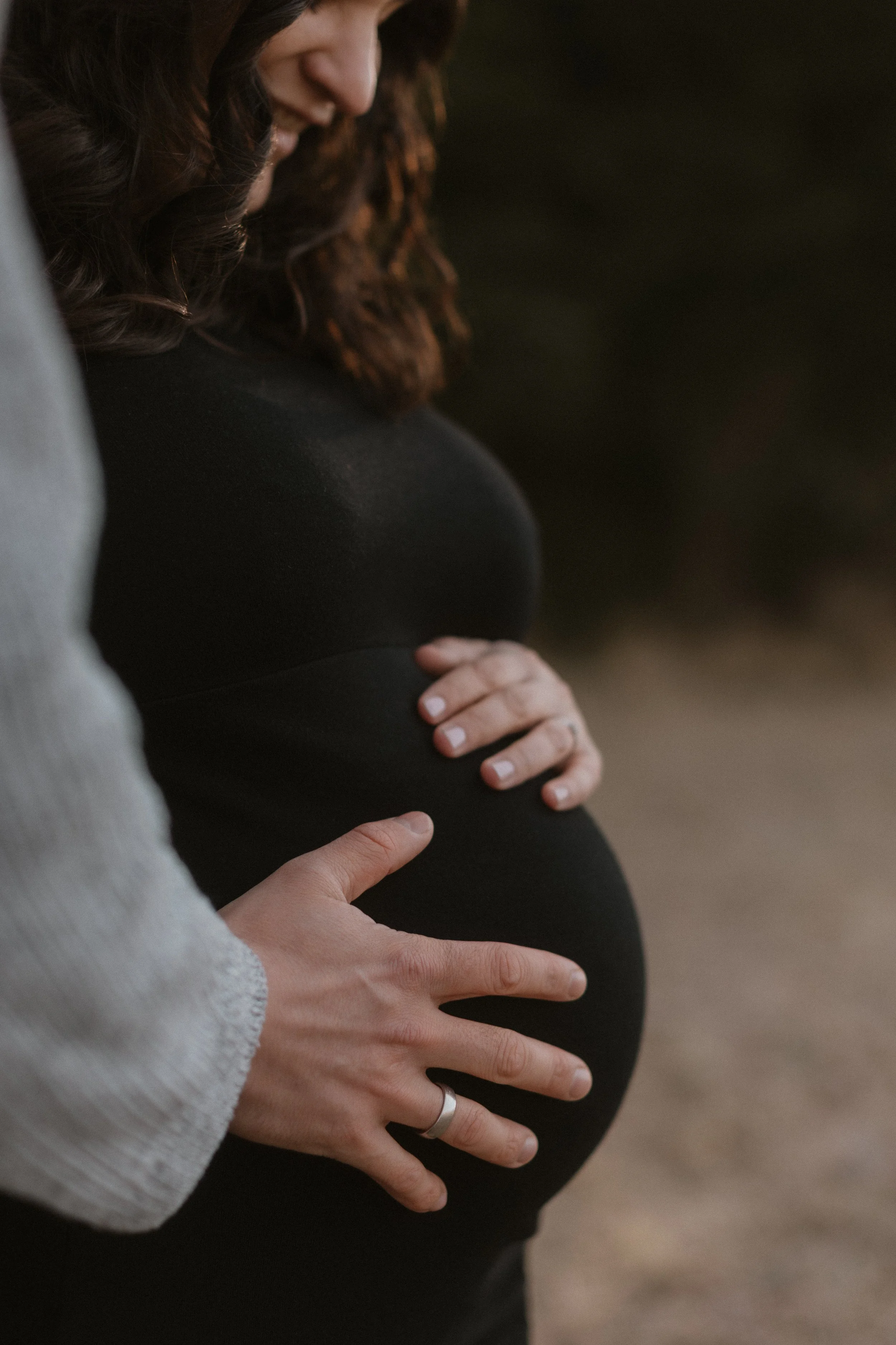 Nahaufnahme eines schwangeren Frauenbauchs, gehalten von einer Person mit einem Ehering auf dem Finger, im Freien bei Sonnenuntergang.