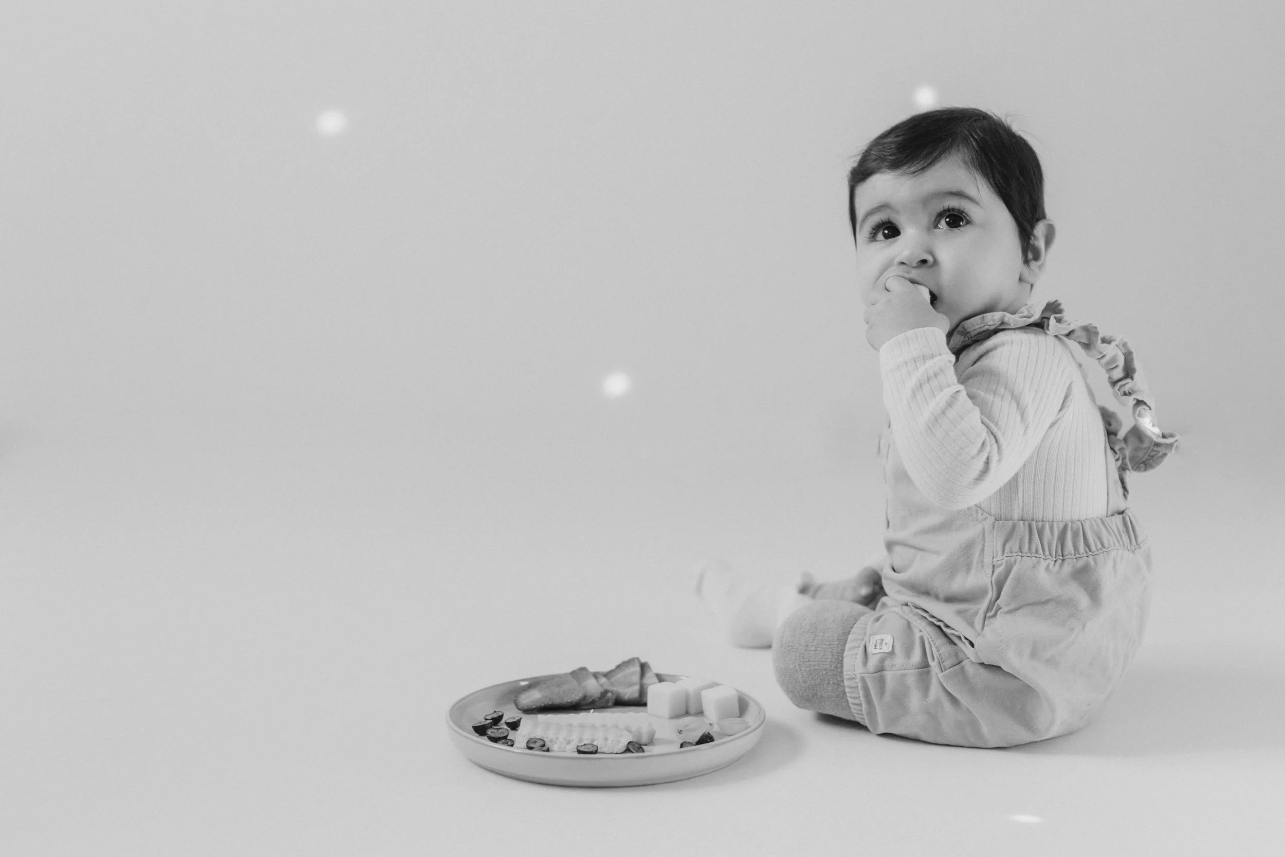 Ein kleiner Junge sitzt auf dem Boden und isst von einem Teller mit verschiedenen Snacks, während er in die Kamera schaut.