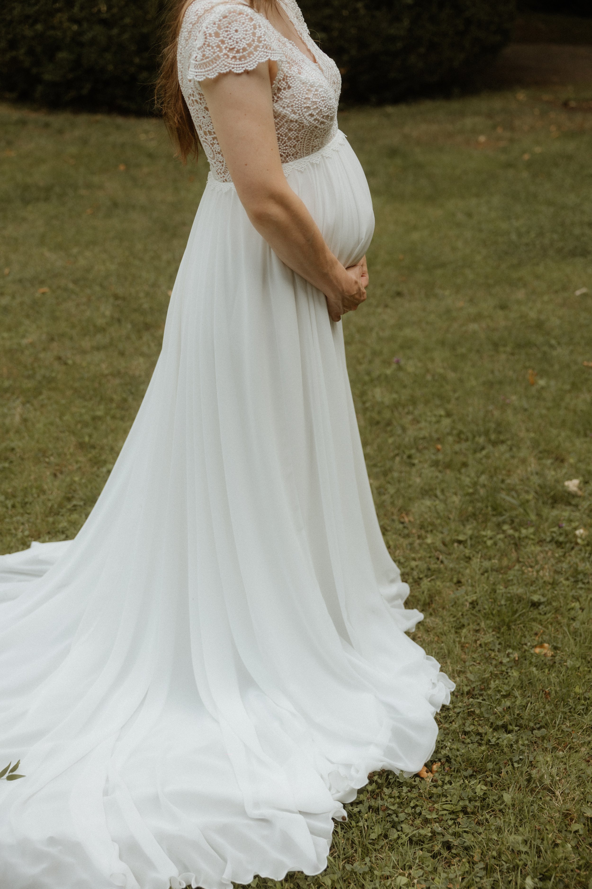 Eine schwangere Frau trägt ein weißes, fließendes Hochzeitskleid mit Spitze und posiert im Freien auf einer Wiese. Fotografiert im Zollernalbkreis von Pia Lydia Heim.