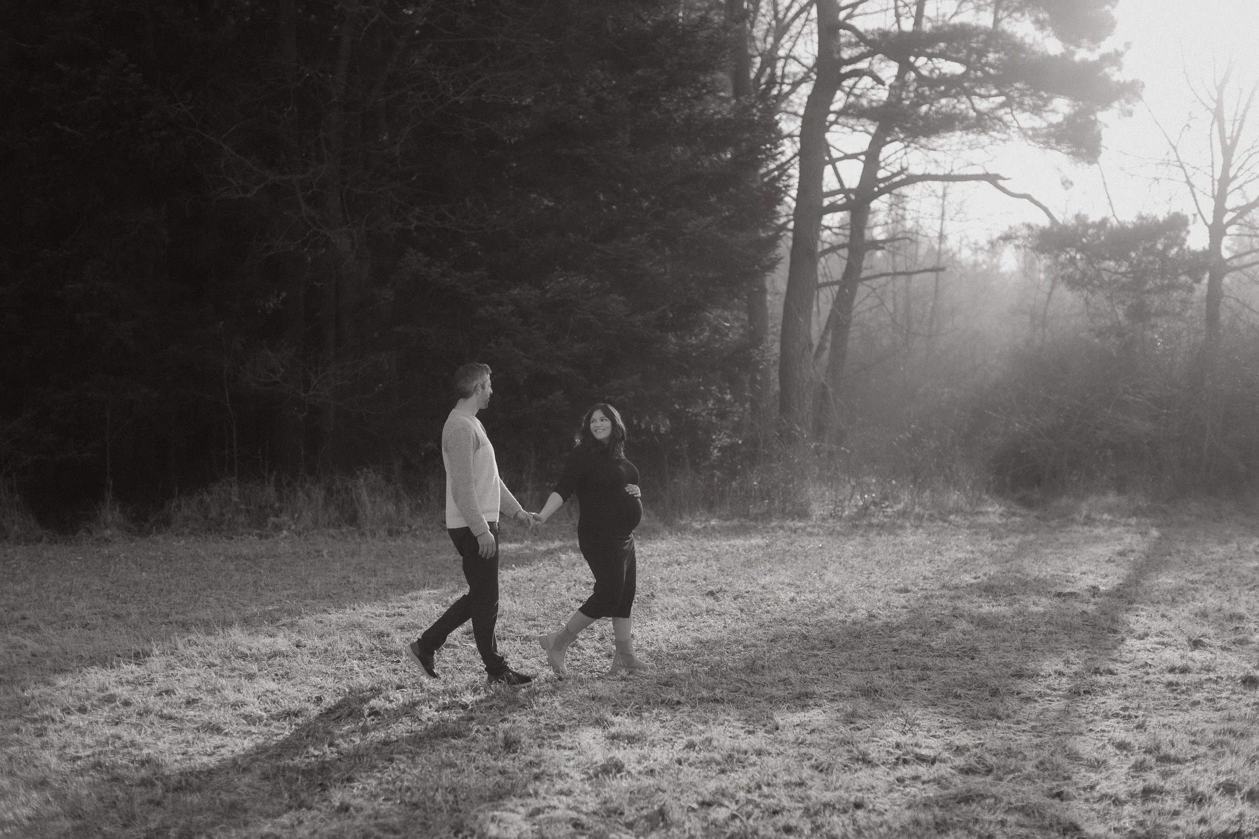 Ein Paar spaziert nackt im Freien, die Frau ist schwanger, umgeben von Bäumen bei Sonnenuntergang.