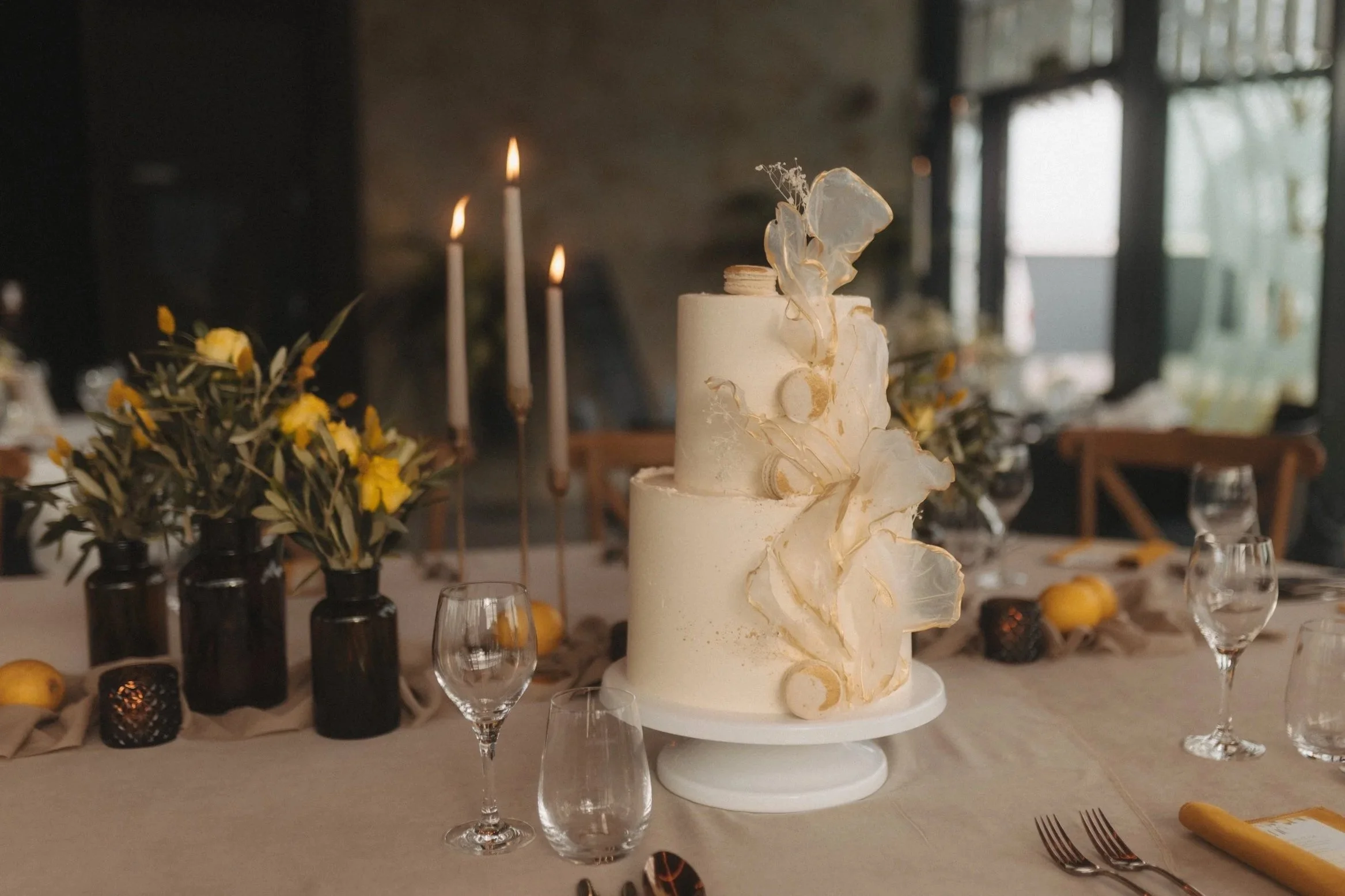 Ein mehrstöckiger Hochzeitstorte der Löwenpatisserie mit weißen und goldenen Dekorationen auf einem festlich gedeckten Tisch mit Gläsern, Blumen und Kerzen in einem eleganten Raum. Von dem Hochzeitsfotograf Pia Heim in der Eventschneiderei.