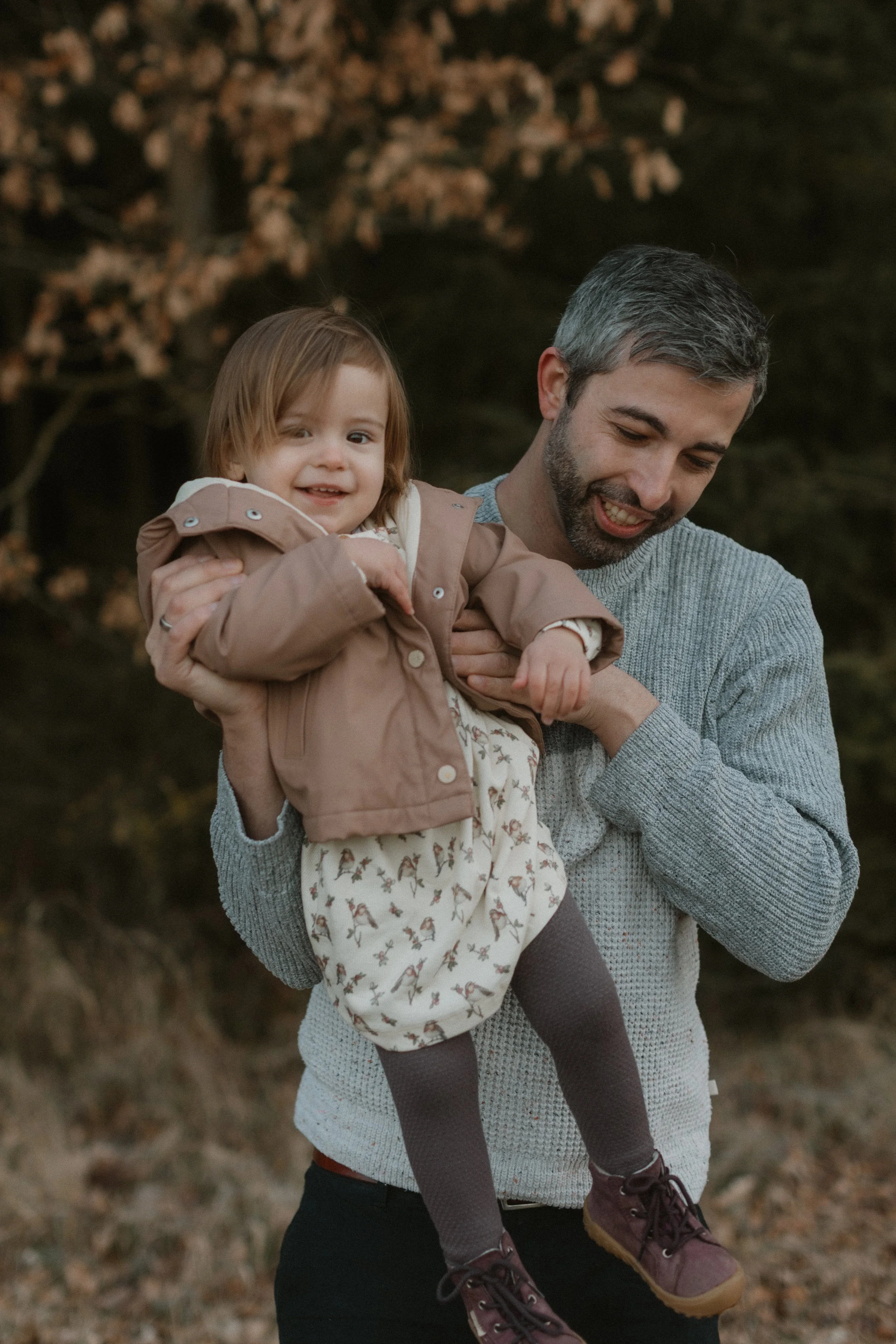 Ein Mann trägt ein kleines Mädchen auf dem Arm in einem herbstlichen Wald, beide lachen und genießen die Natur.