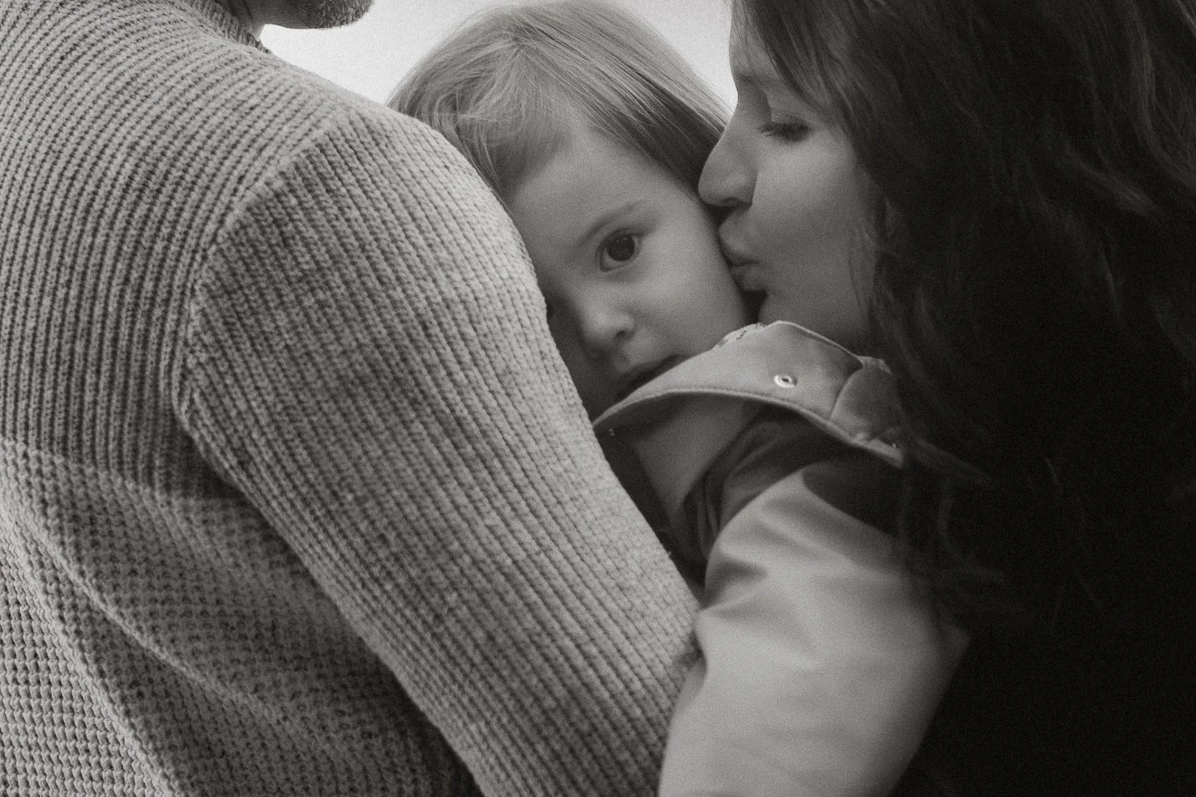 Eine Frau küsst ein kleines Kind, das in ihre Richtung schaut und in den Arm einer Person mit hellem Pullover liegt, im Schwarz-Weiß.