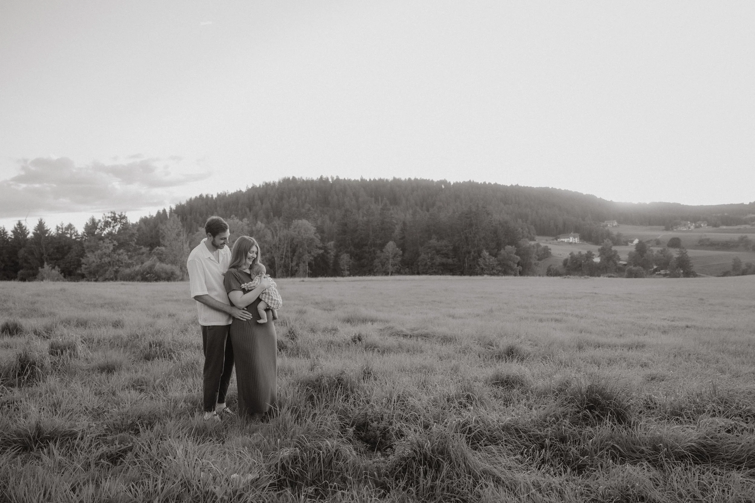 Ein Paar hält ein schlafendes Baby in einem offenen Grasfeld, umgeben von Bergen und einer ländlichen Landschaft, in Schwarz-Weiß aufgenommen.