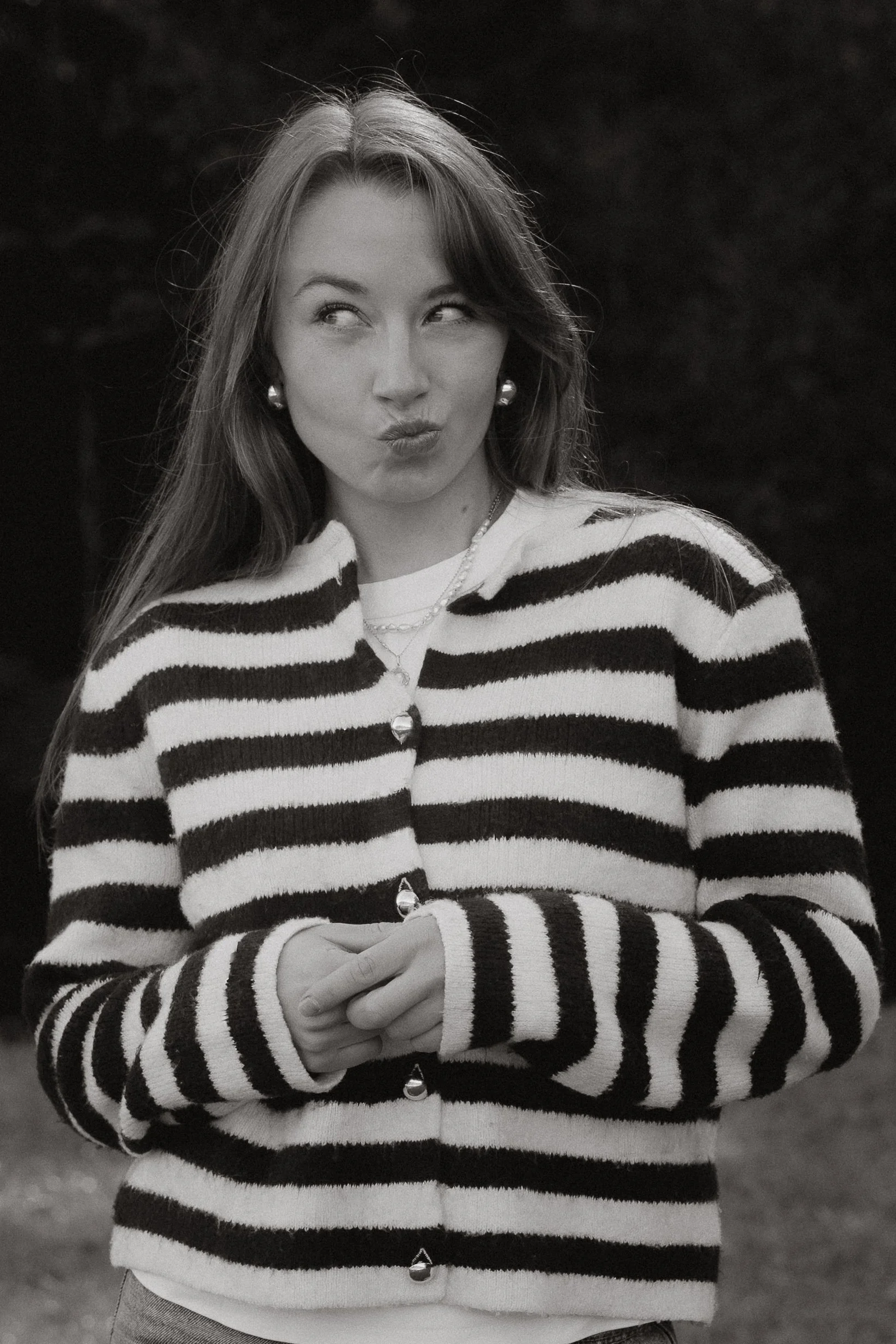 Schwarz-weiße gestreifte Strickjacke, lächelnde junge Frau mit langen Haaren und Ohrringen, die nach oben schaut und die Lippen schürzt. In einer natürlichen Umgebung mit dunklem Hintergrund.