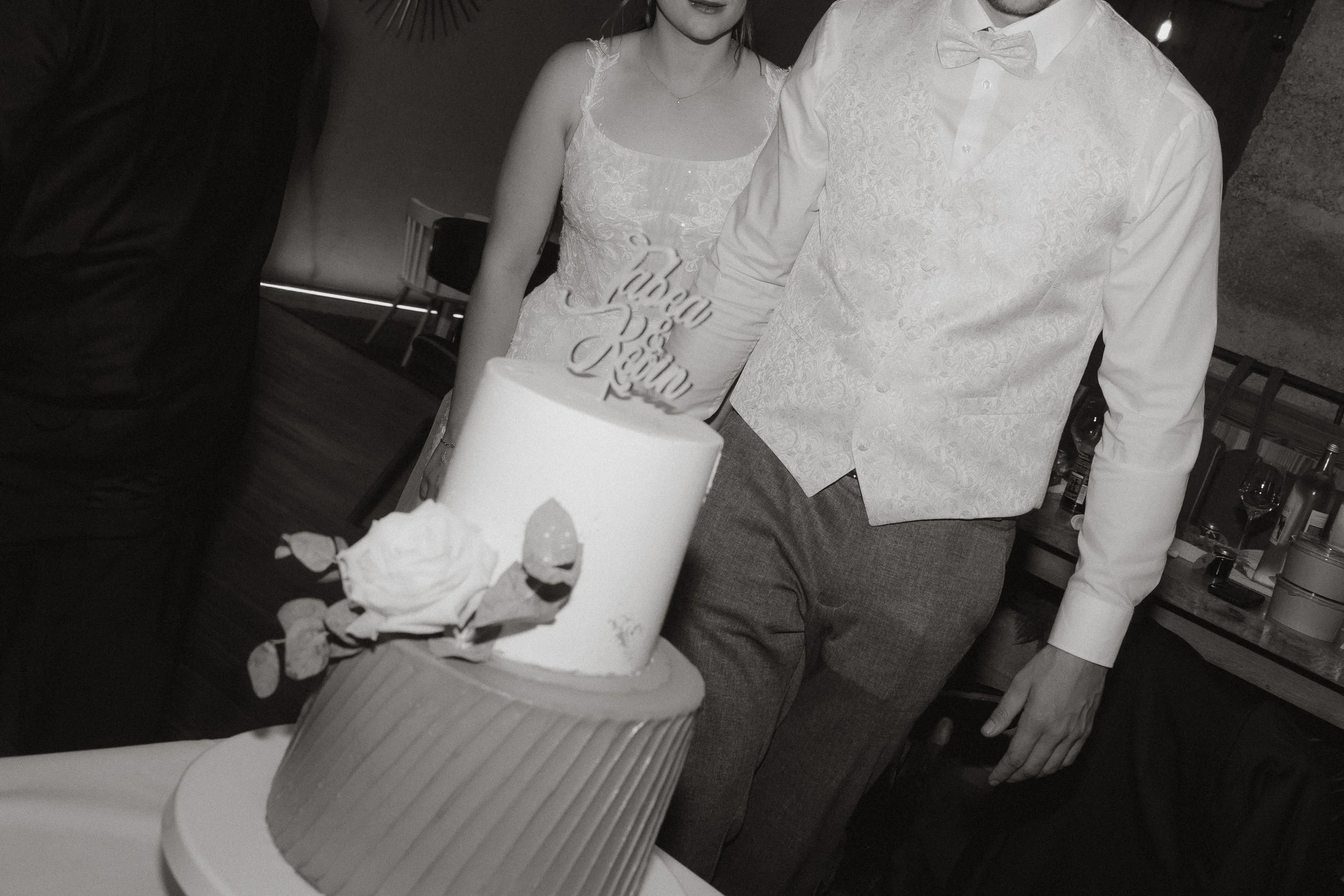 Eine Hochzeitstorte von der Löwenpatisserie, Schönwald mit einem Topper auf einem Tisch bei einer Hochzeitsfeier. Im Hintergrund sind zwei Personen in Hochzeitskleidung. Aufgenommen in der Säge, Niedereschach mit Blitz von Pia Lydia Heim. 