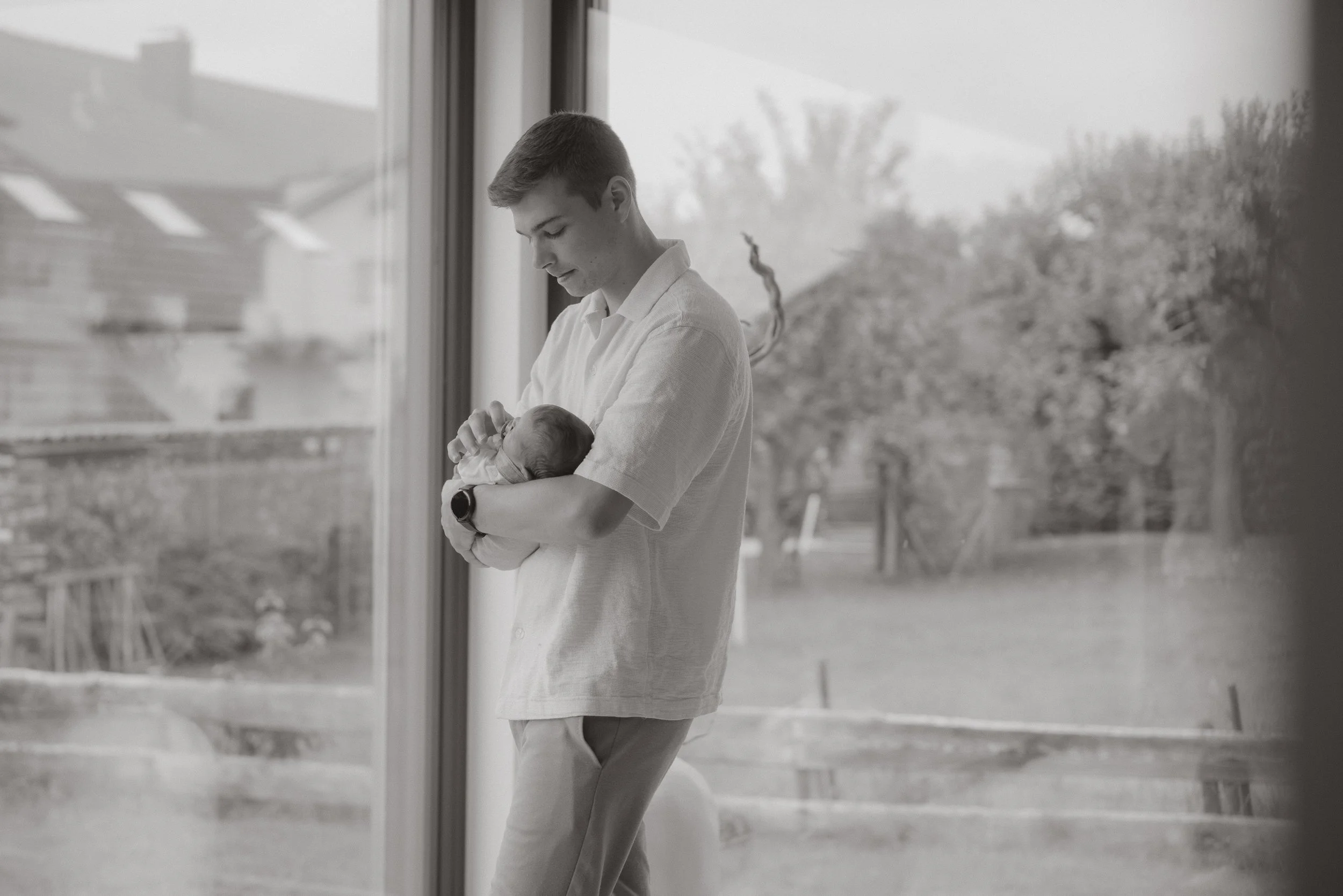 Ein junger Mann hält ein neugeborenes Baby in den Armen, steht neben einem Fenster in einem Haus, im Hintergrund sind Bäume und ein Garten zu sehen.