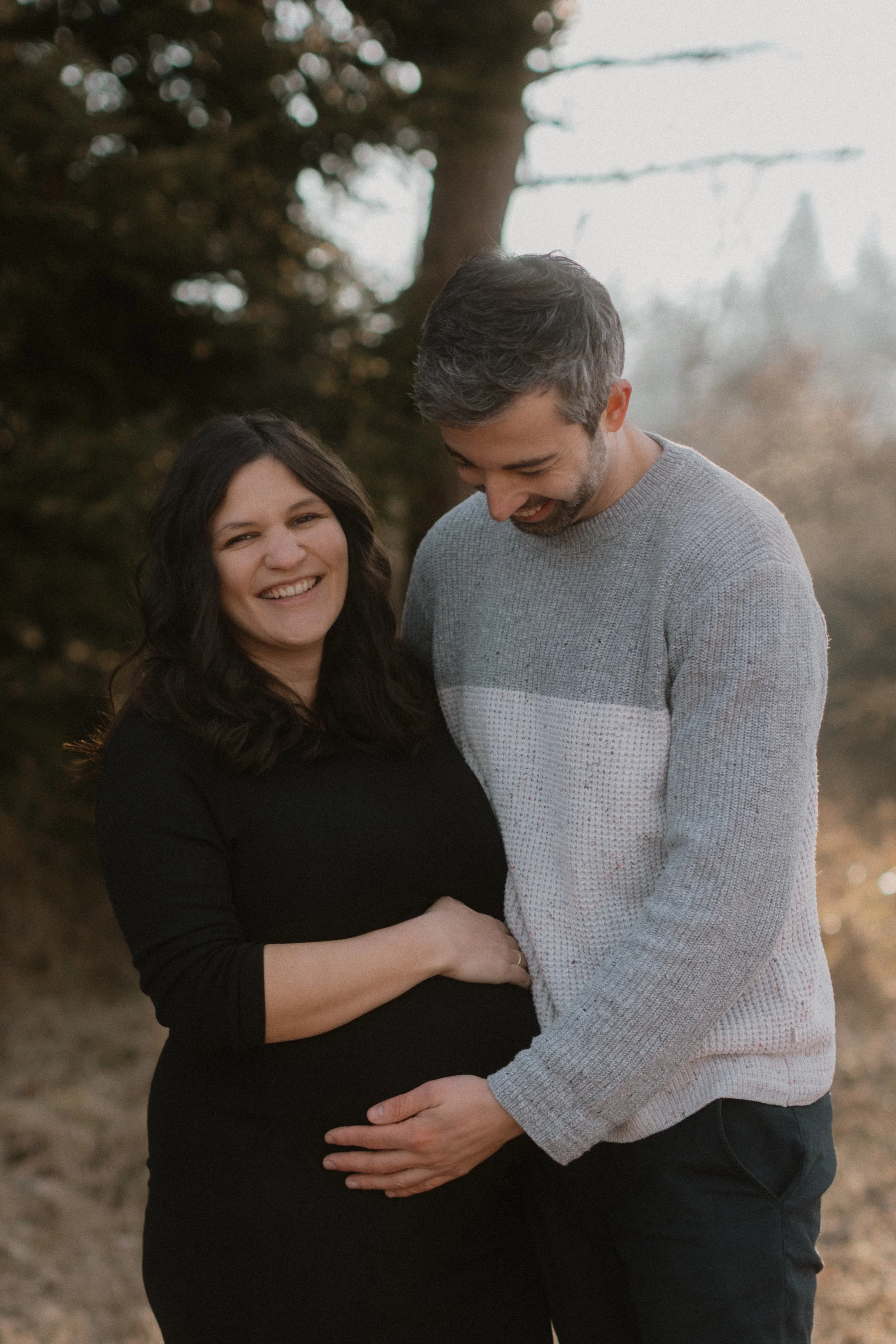 Ein glückliches schwangeres Paar umarmt sich im Freien, ein Mann trägt einen grauen Pullover und eine Frau ein schwarzes Kleid, beide lächeln und schauen zueinander.
