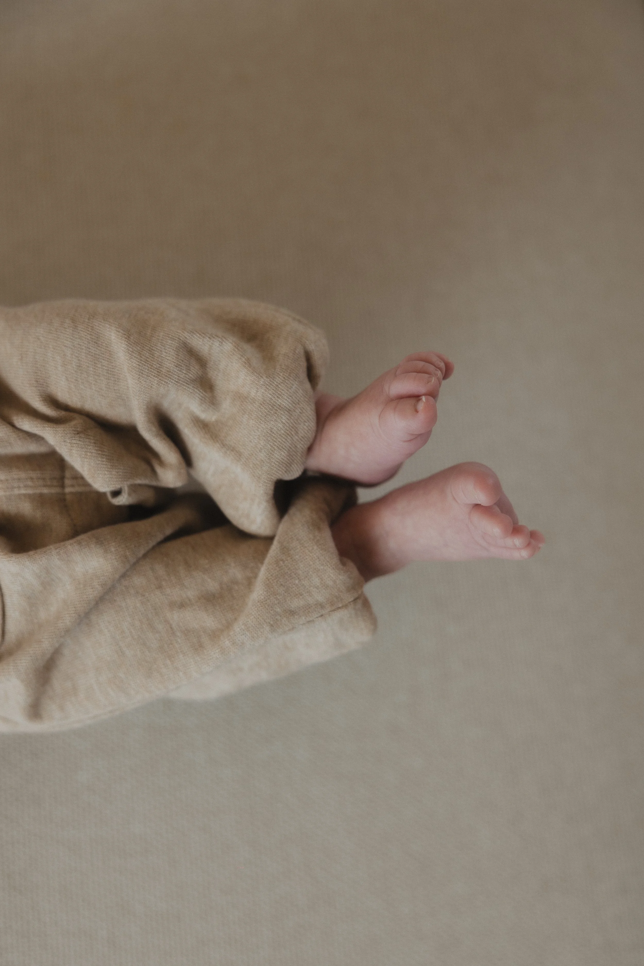 Nahaufnahme von kleinen Babyfüßen in einem bodenlangen, beige Stoffoverall, vor neutralem Hintergrund.