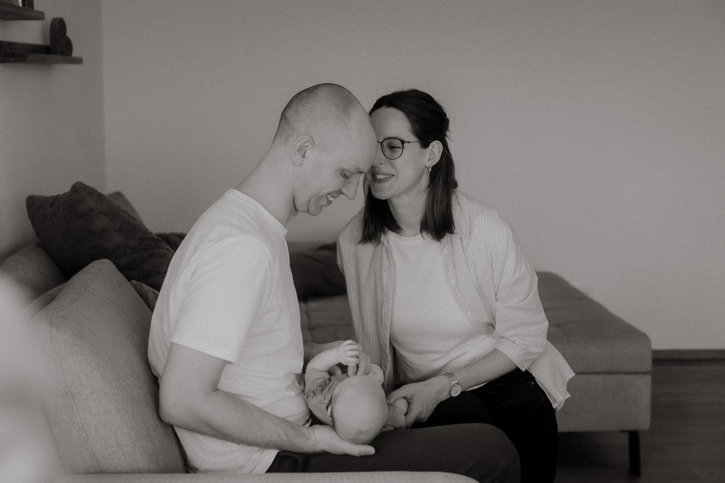 Ein Mann sitzt auf einer Couch mit einem Baby in den Armen, während eine Frau neben ihm sitzt, beide lächeln.