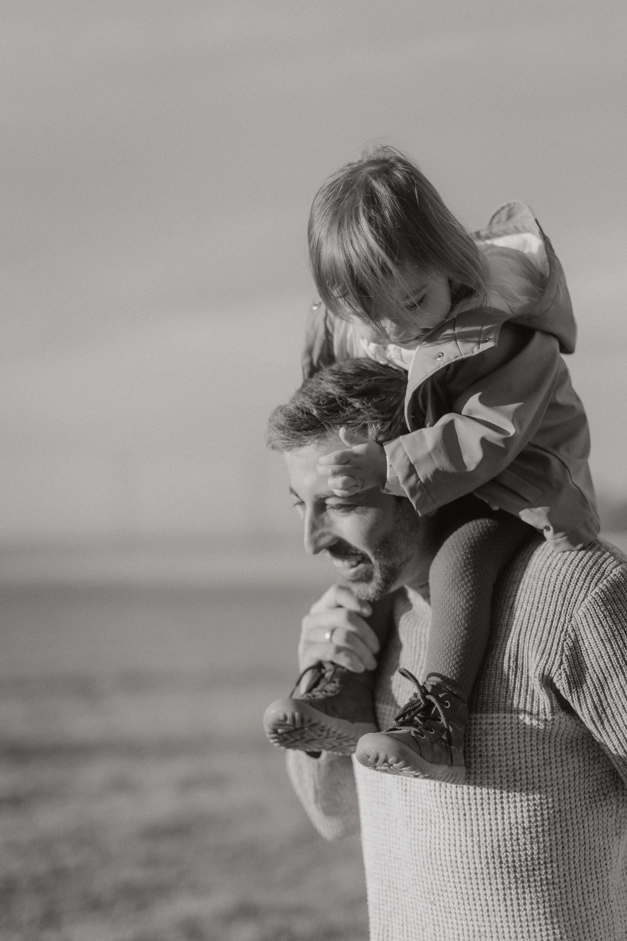 Ein Mann trägt ein Mädchen auf den Schultern. Das Mädchen sitzt auf den Schultern und hält den Kopf des Mannes fest. Das Bild ist schwarz-weiß.