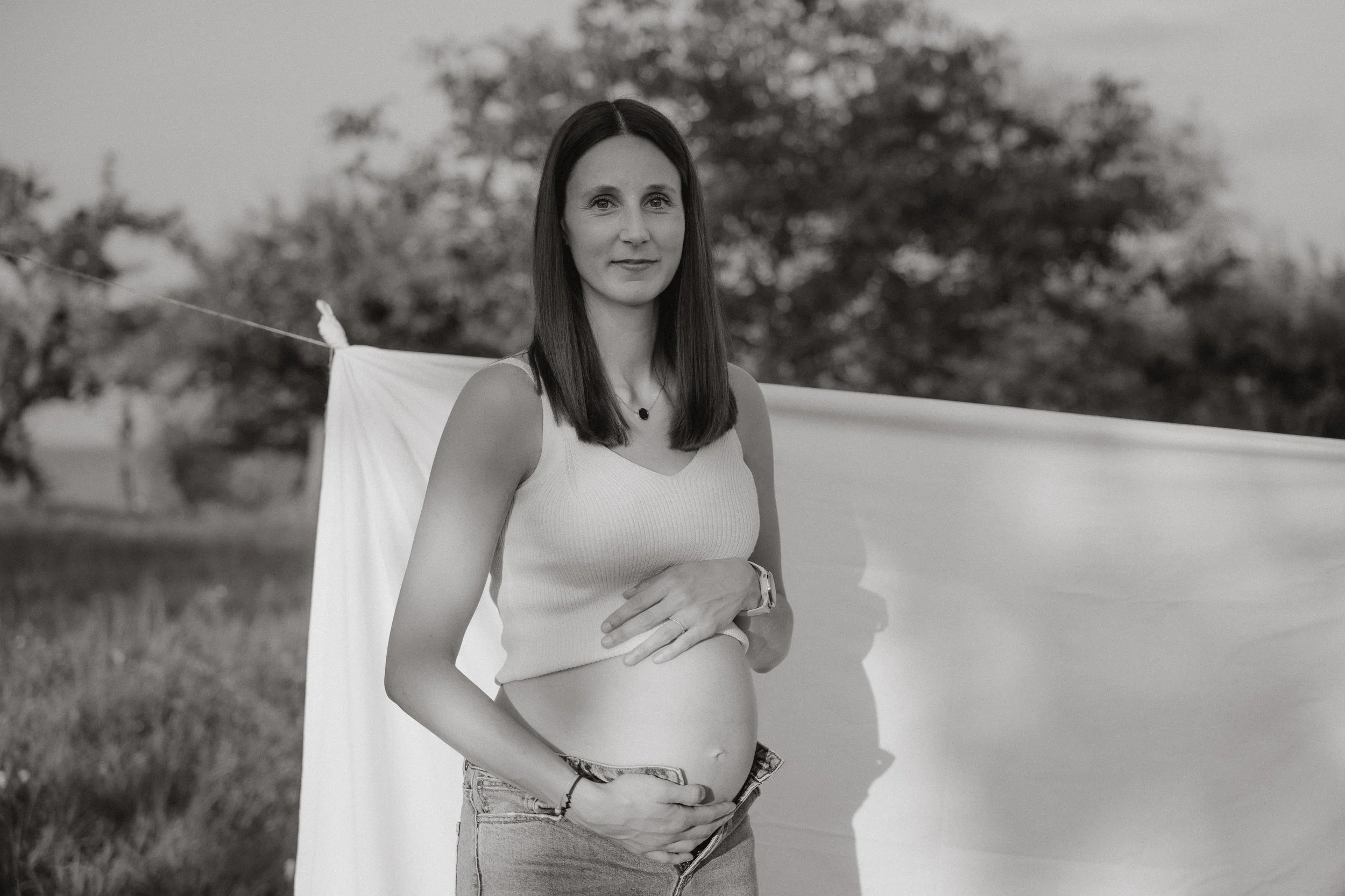 Schwangere Frau in Top und Jeans steht in der Natur vor einer weißen Leinwand bei ihrem Babybauchshooting.