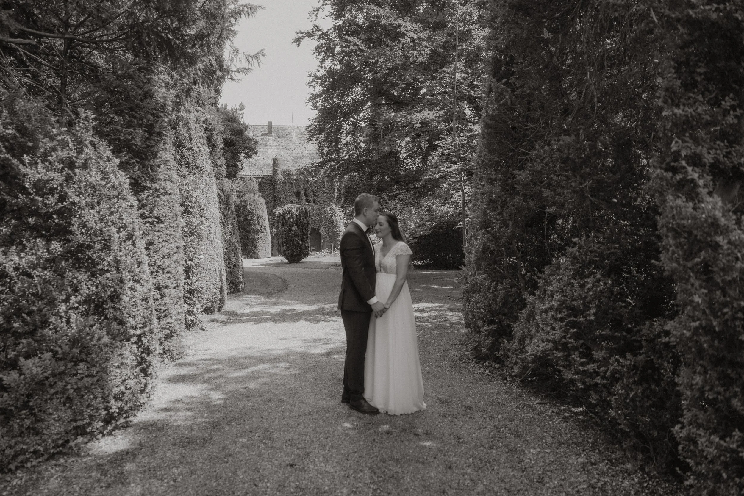 Ein Brautpaar steht in einem Garten, von Bäumen und Hecken umgeben, bei Sonnenlicht.
