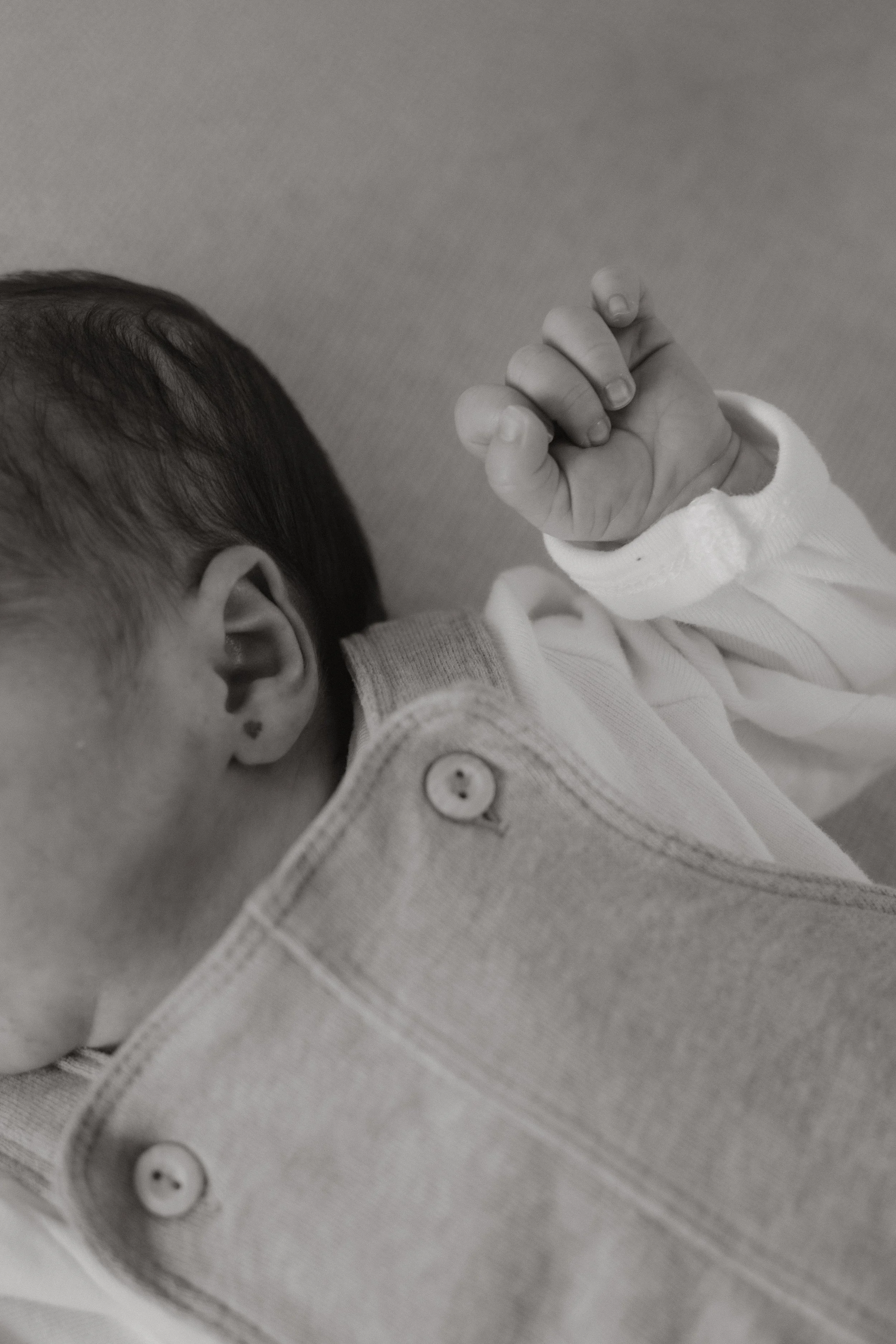 Schwarz-weiß-Foto eines Neugeborenen mit geschlossenem Ohr und einer Hand, die eine Faust macht, in einem weißen Kleid. Der Hintergrund ist neutral.