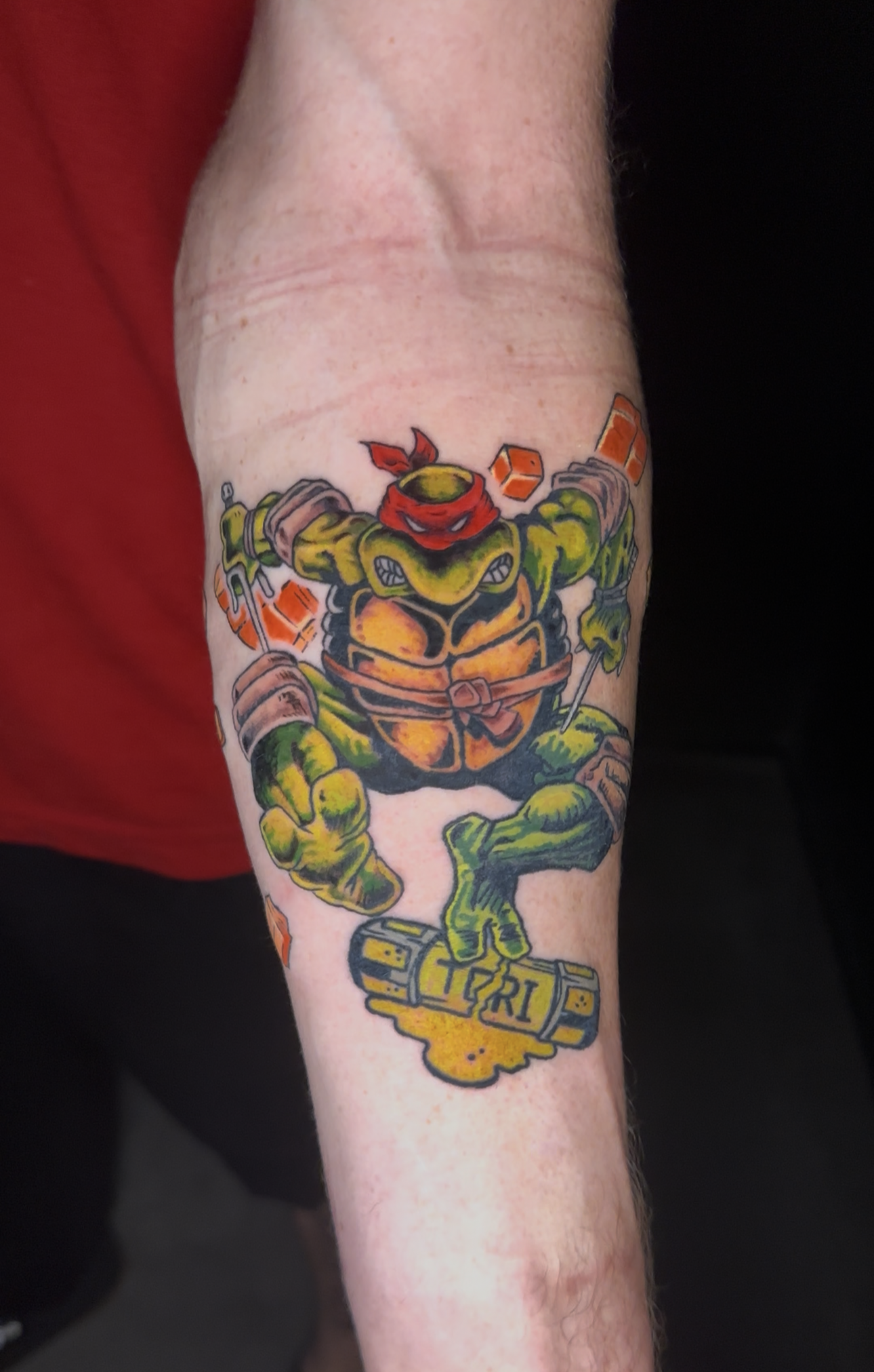 Colorful tattoo of a Teenage Mutant Ninja Turtle character on a person's arm, wearing a red mask, holding weapons, with the word 'RI' visible on a piece of armor or a weapon.