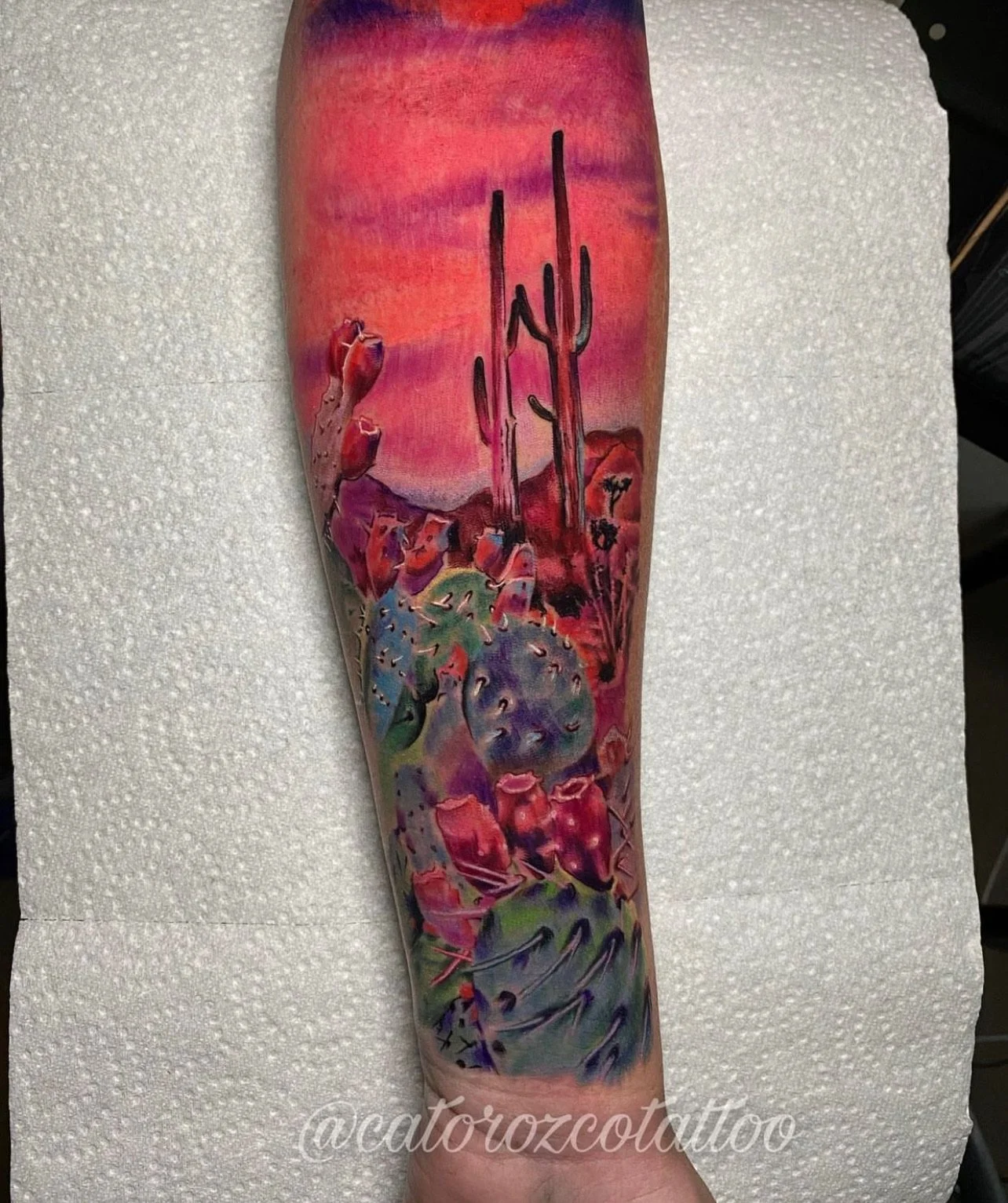 Colorful tattoo of a desert landscape with cacti and mountains on a person's forearm.