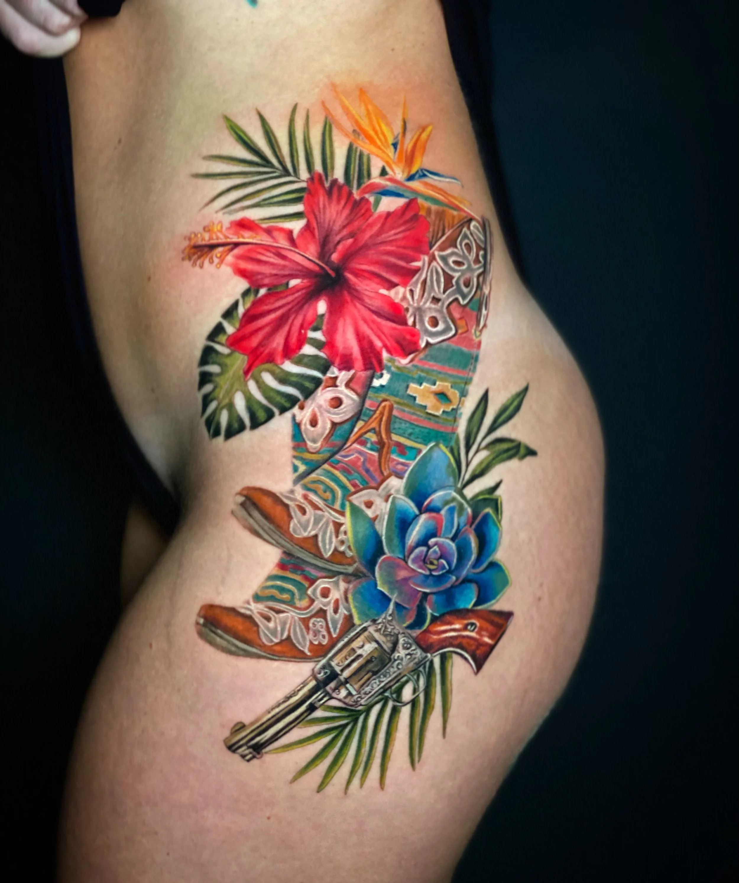 Colorful tattoo featuring tropical elements, including a red hibiscus flower, orange and blue bird of paradise flowers, a butterfly, green palm leaves, and tribal patterns, all on a person's arm.