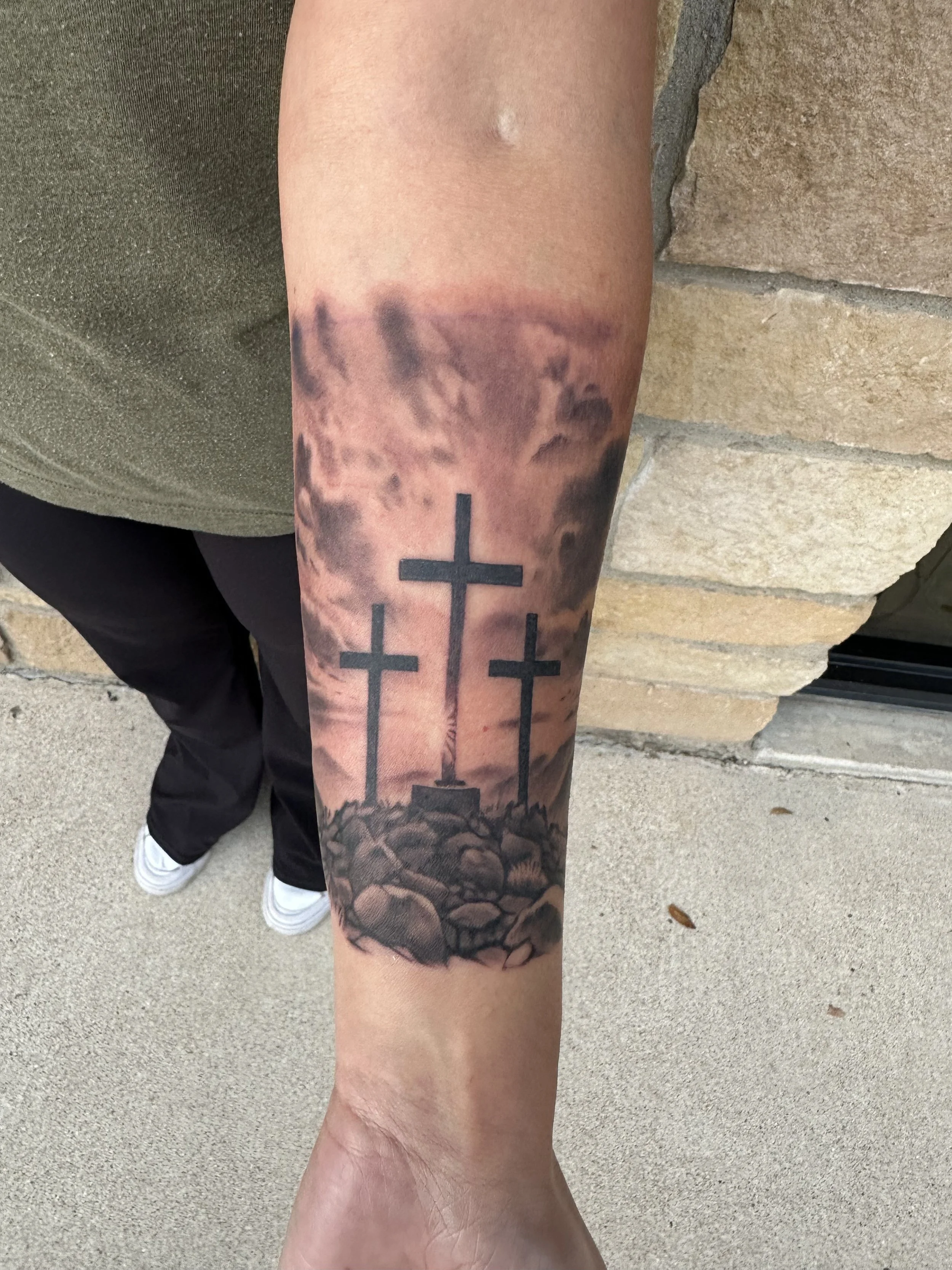 Tattoo of three crosses on rocky ground against a cloudy sky on a person's forearm.