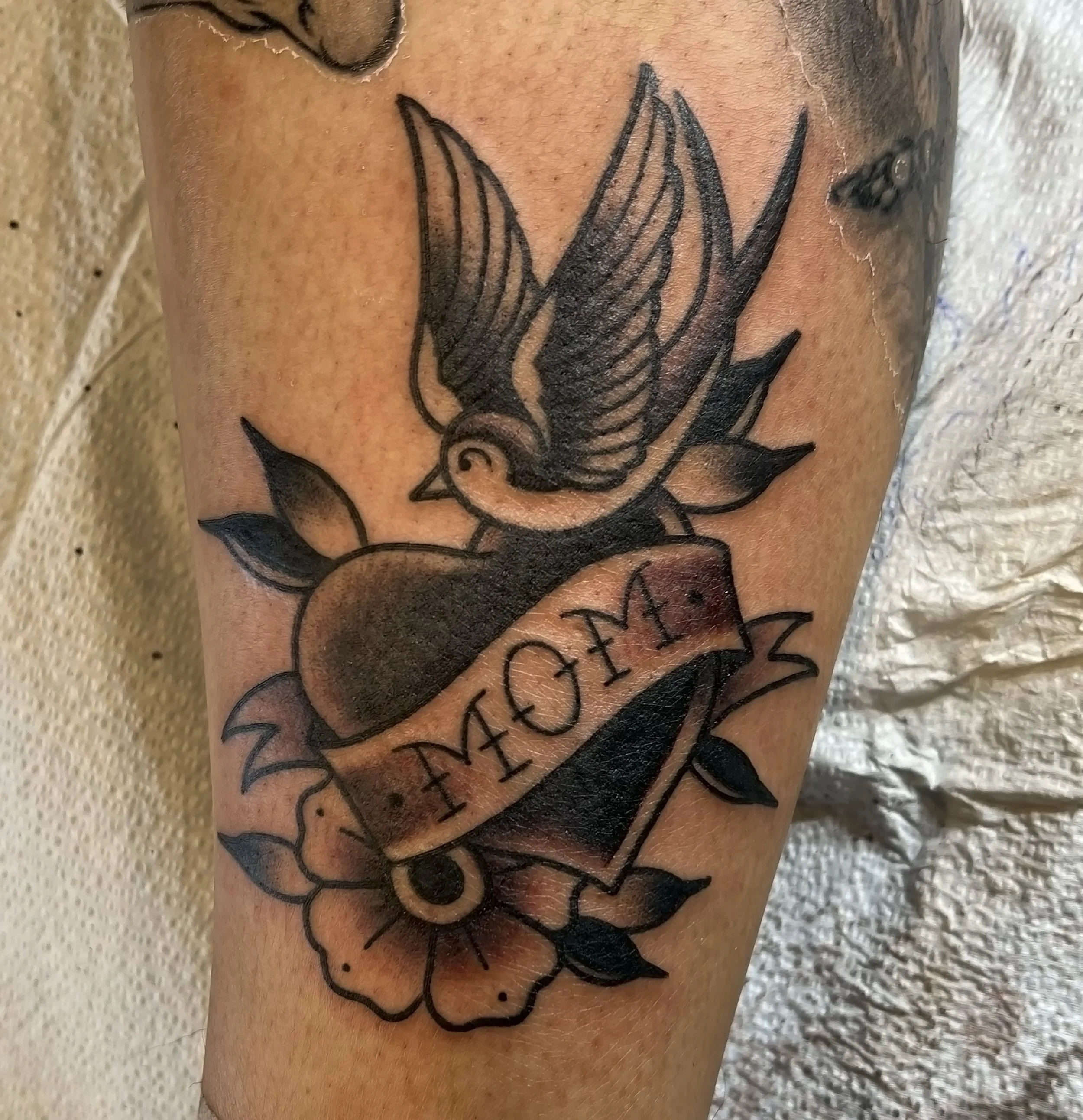 a black. and grey traditional style tattoo of a heart with a banner that says "mom" over it, with a sparrow on top and a flower and leaves surrounding the heart