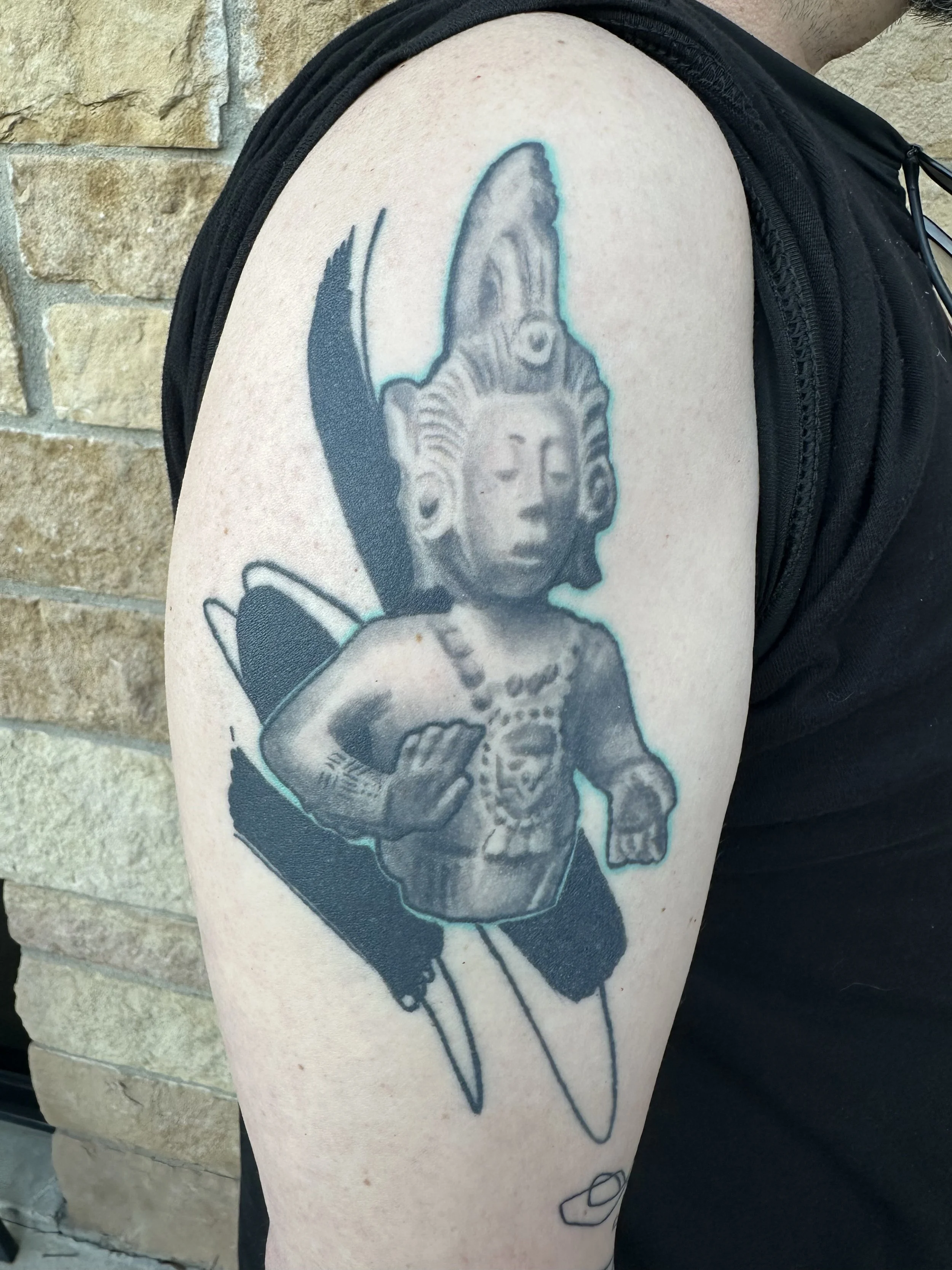 Tattoo of a fairy with a humanoid face, butterfly wings, wearing a beaded necklace, with a knife and a small object beneath.