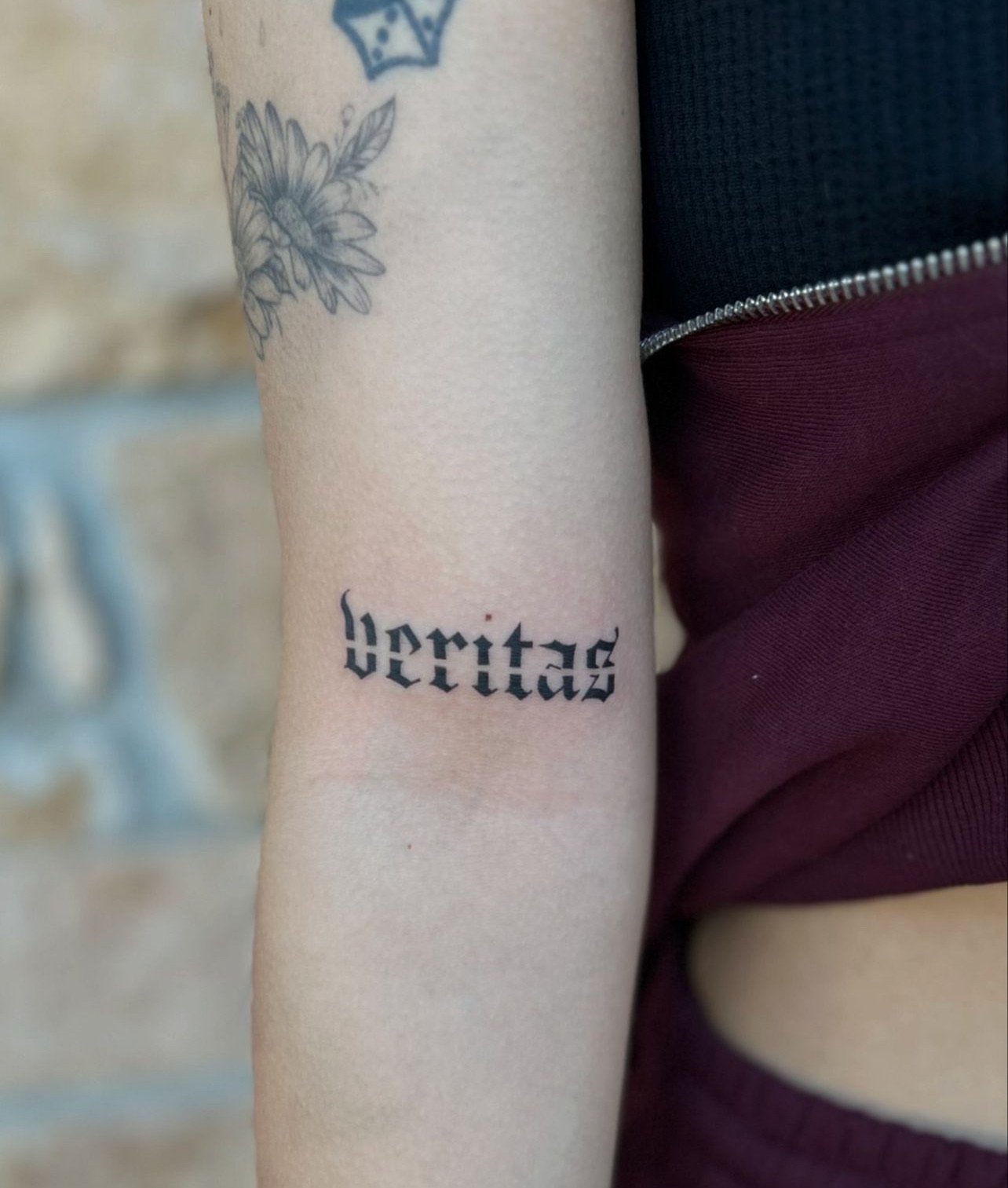 Tattoo of the word 'veritas' in blackletter font on a person's forearm, with a flower tattoo partially visible above.