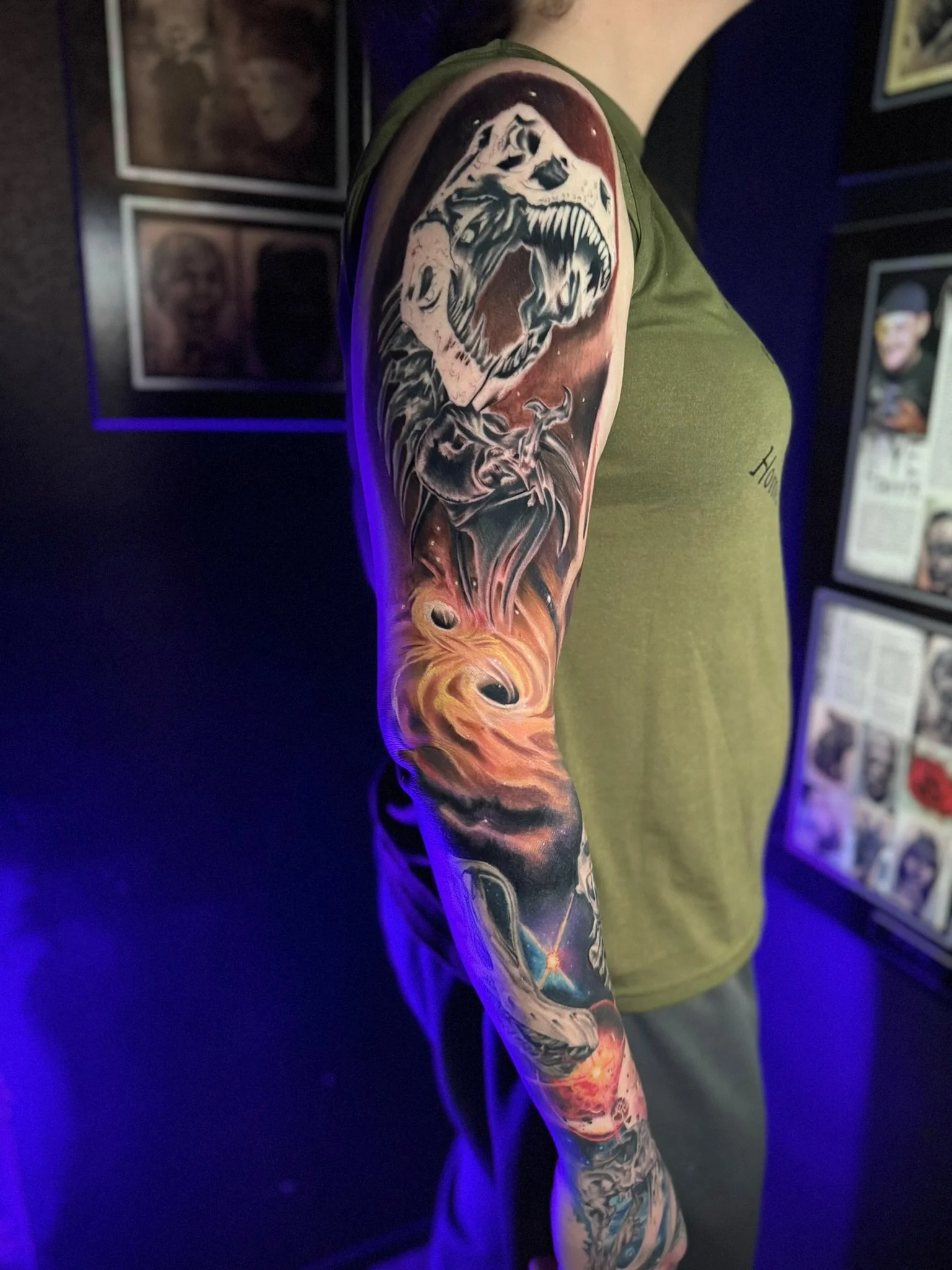 Tattoo of a roaring dinosaur head and a cosmic scene on a person's upper arm.