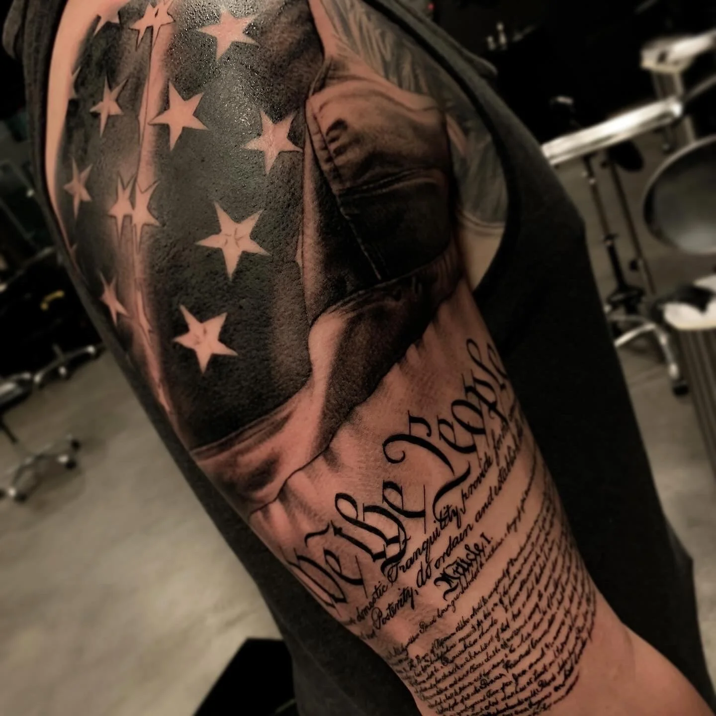 Close-up of a person's upper arm and shoulder displaying a large tattoo of an American flag with stars and stripes, accompanied by script writing and text below.