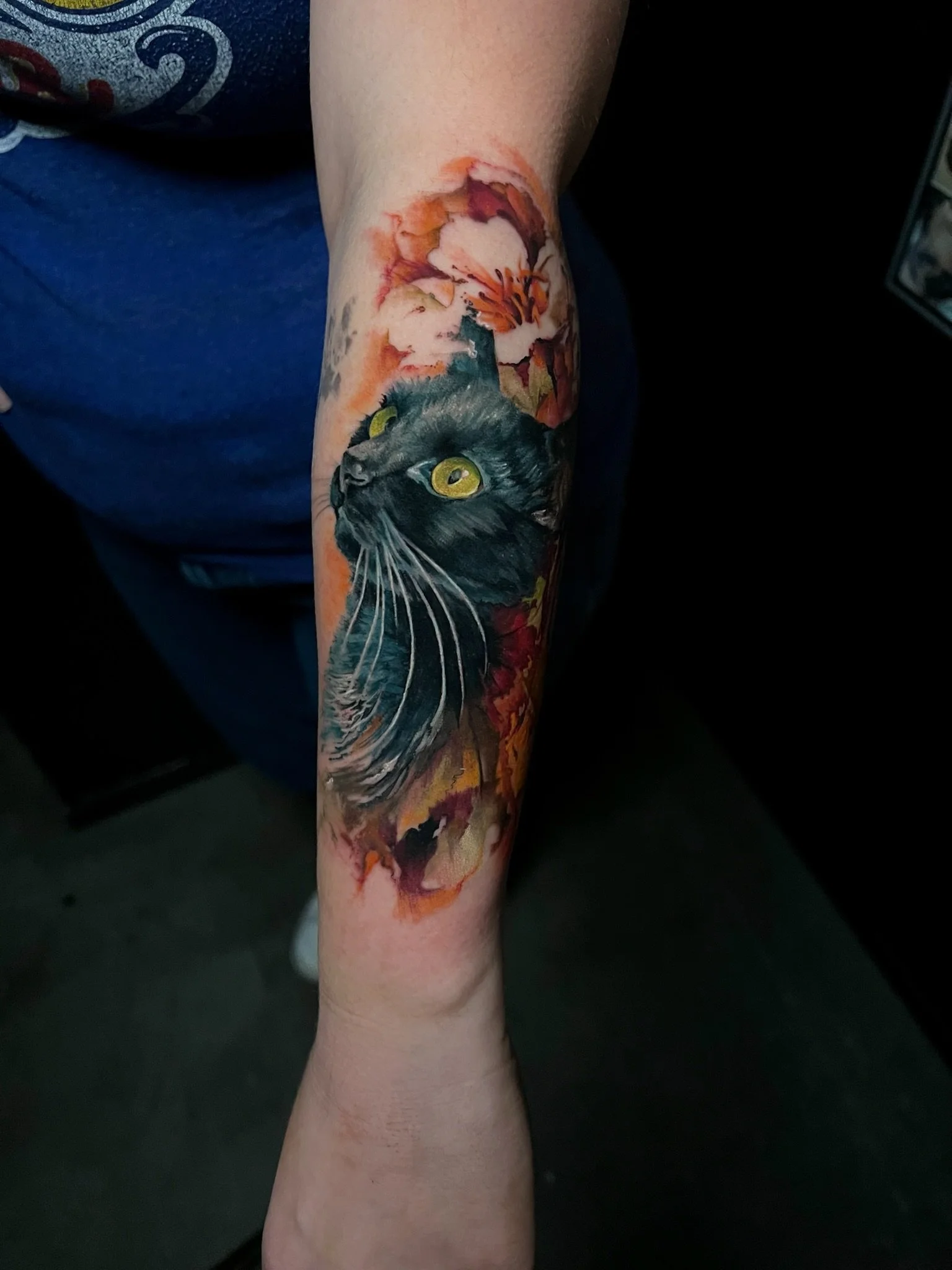 Colorful tattoo of a black cat with yellow eyes surrounded by orange and pink flowers.