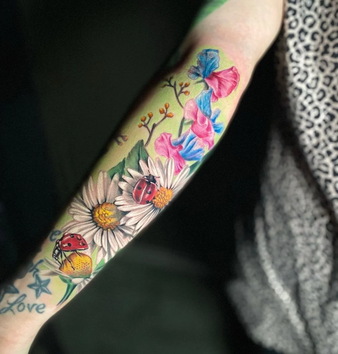 Tattoo of a floral design with pink, blue, and purple flowers, two ladybugs on white daisies, and the word 'Love' written in blue at the bottom.