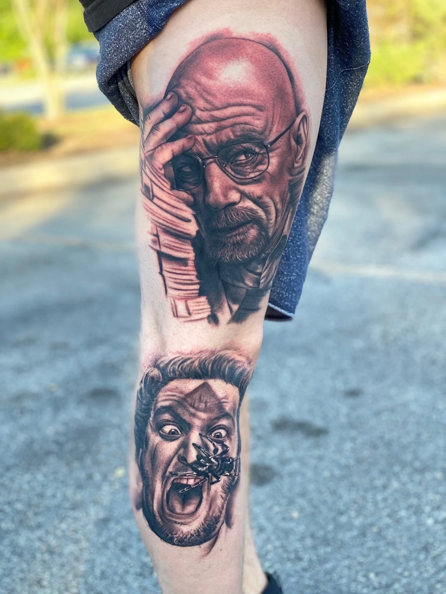Tattoo of two faces on a person's leg. The upper face is an older man with glasses, bald head, and beard, holding his forehead. The lower face is a man with short hair and beard, with an angry expression and a large insect on his tongue.