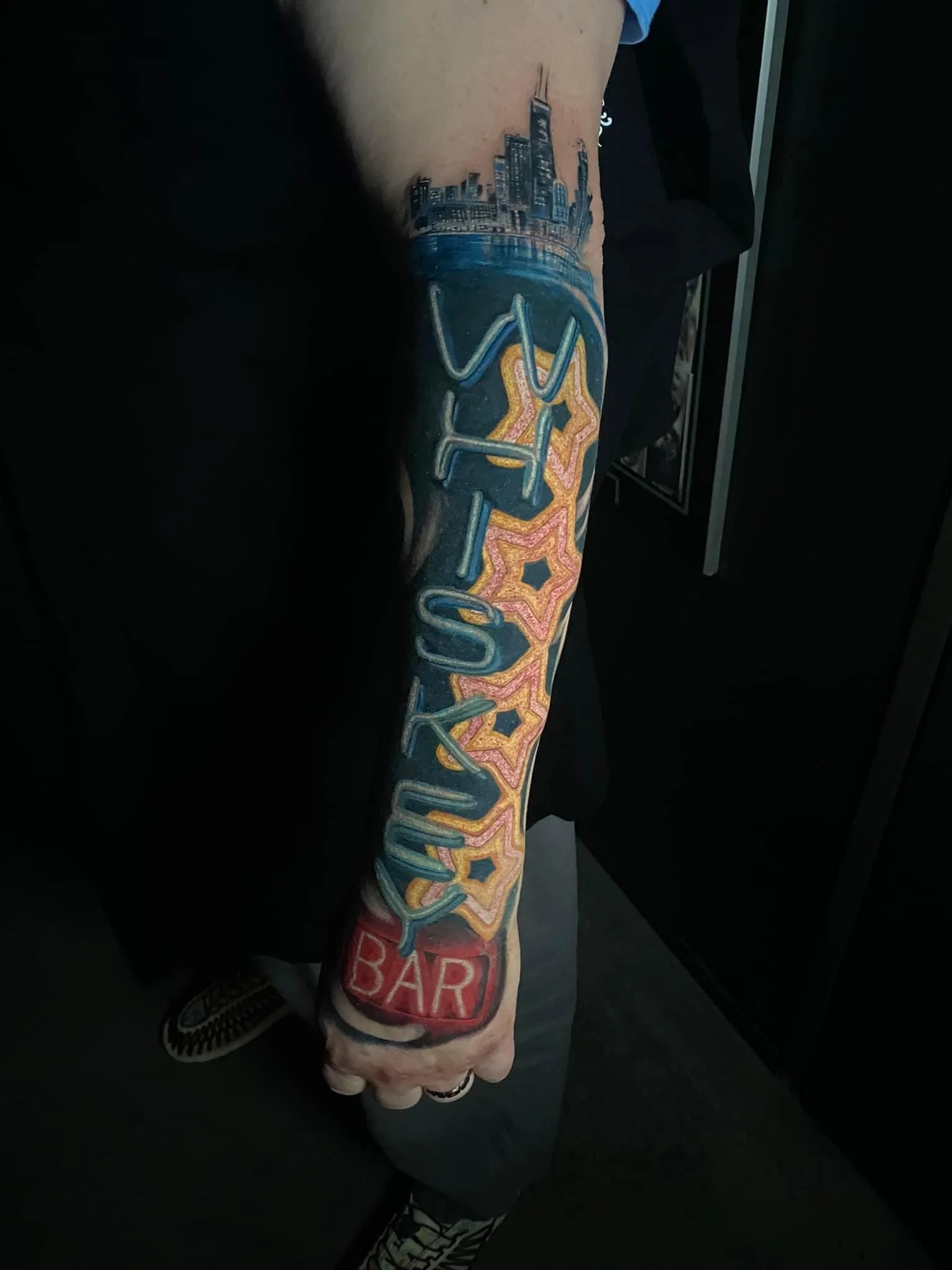 Colorful tattoo on a person's forearm with the words 'NYC' and 'Bar', featuring cityscape and stars.