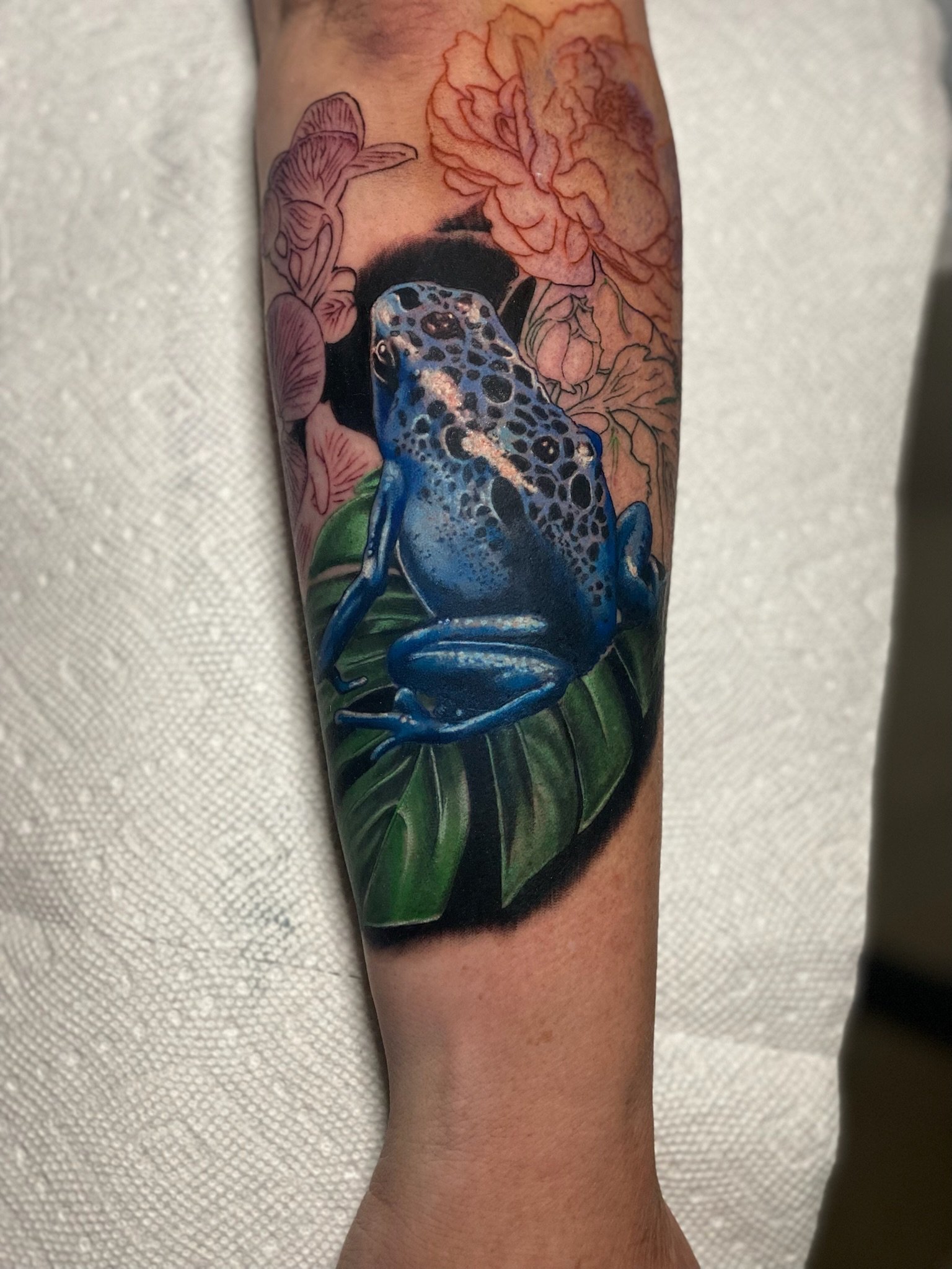 Tattoo of a blue leopard frog sitting on a large green leaf, surrounded by pink and orange flowers.