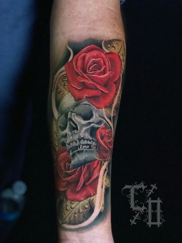 Tattoo of a realistic human skull surrounded by red roses, with gold and green leaves.