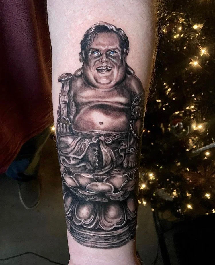 Tattoo of a humorous, realistic-style depiction of a chubby man with light brown hair and bright blue eyes, sitting in a semi-portrait position with an expressive face, on a Buddhist or religious statue base.