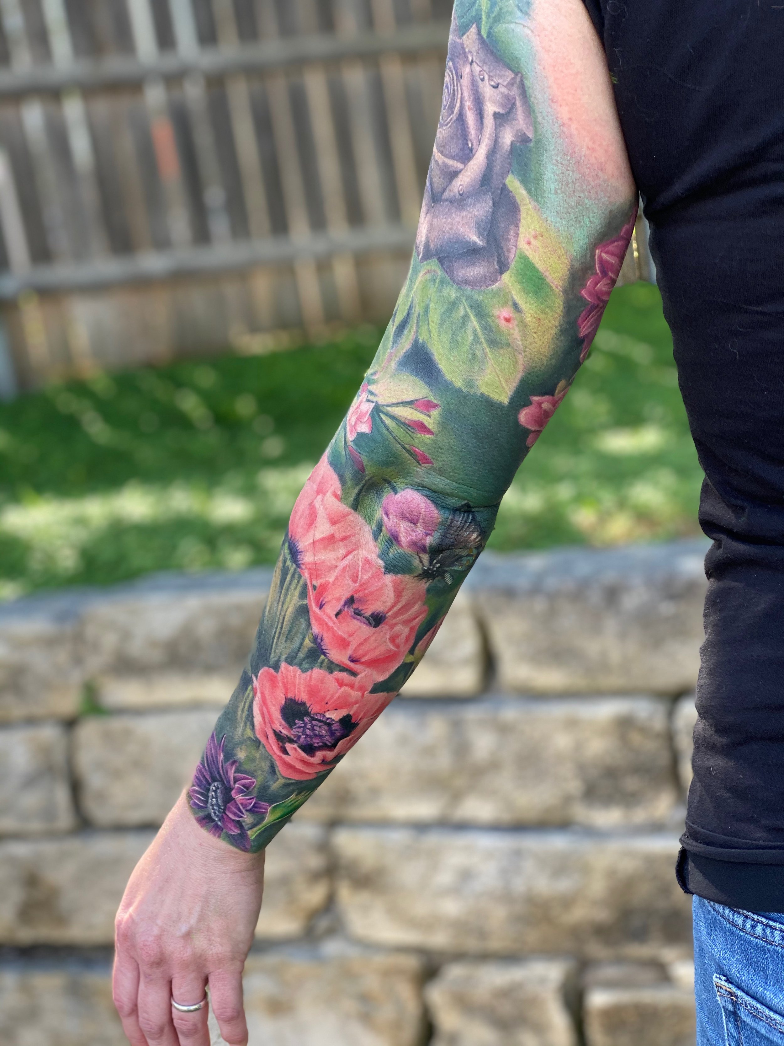 Close-up of two people holding hands, with one person's arm covered in colorful tattoos featuring flowers and a bird, and the other person's hand and arm visible. The background shows a wooden fence and grass.